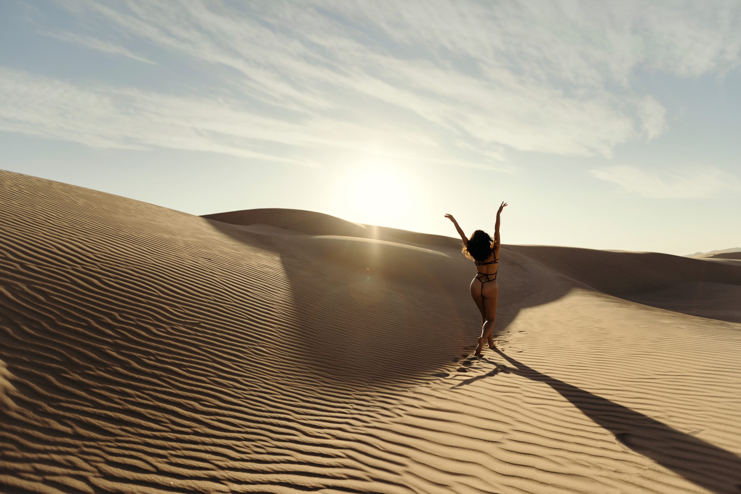 Person standing on sand dunes with arms raised, sun setting behind dunes, long shadow cast on sand, clear sky.