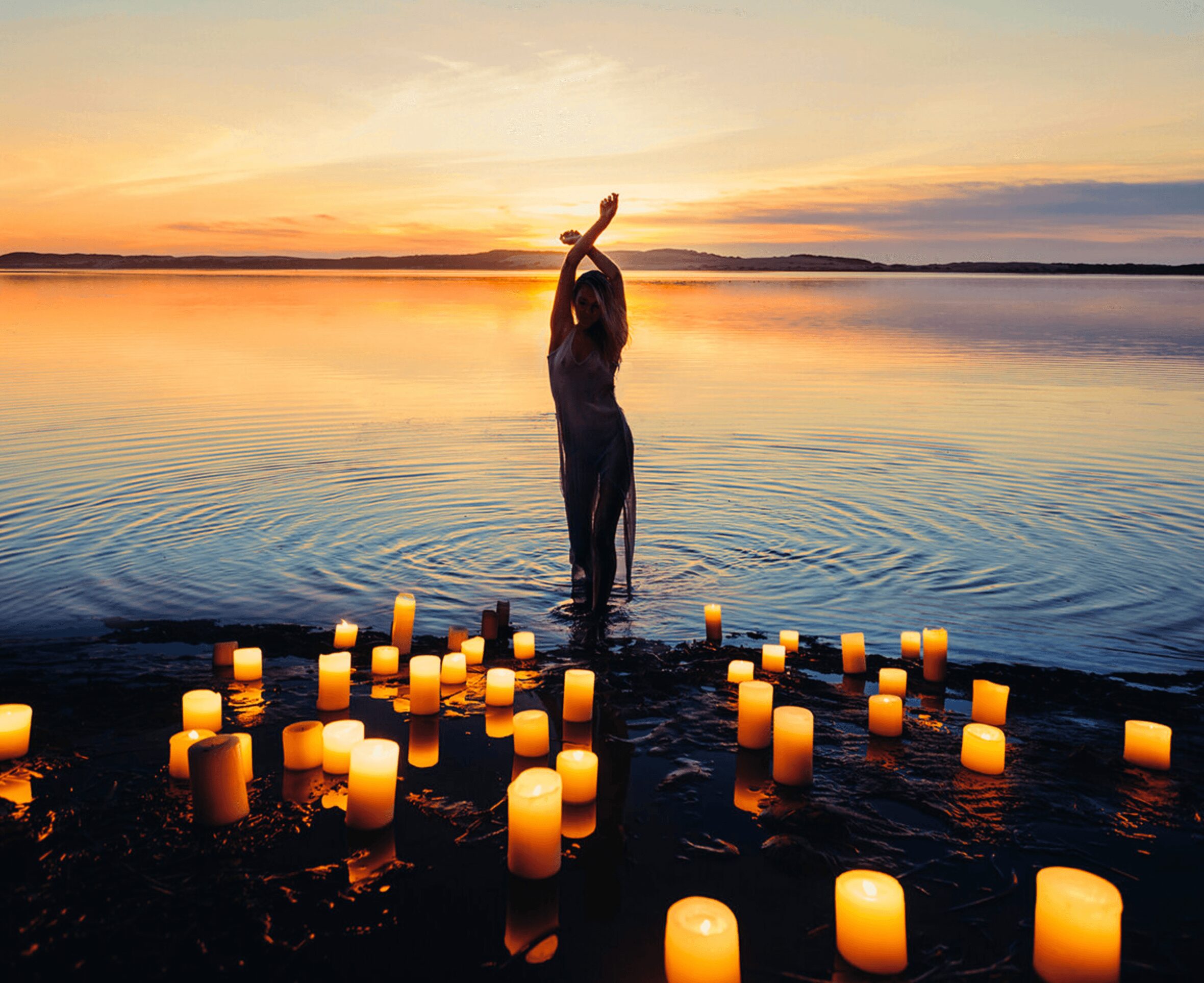 Person standing in water at sunset with candles on the shore, arms raised above head, calm scene.