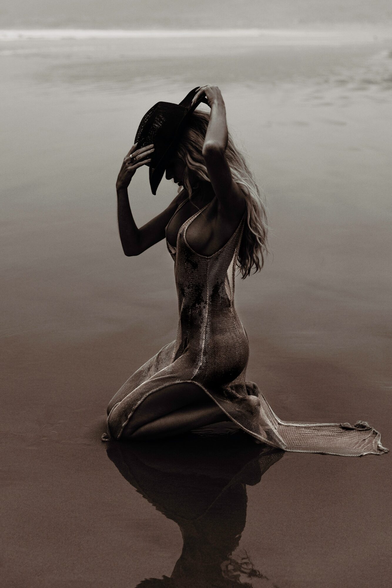 Kneeling woman on beach with long hair, touching her head, water and horizon in background.