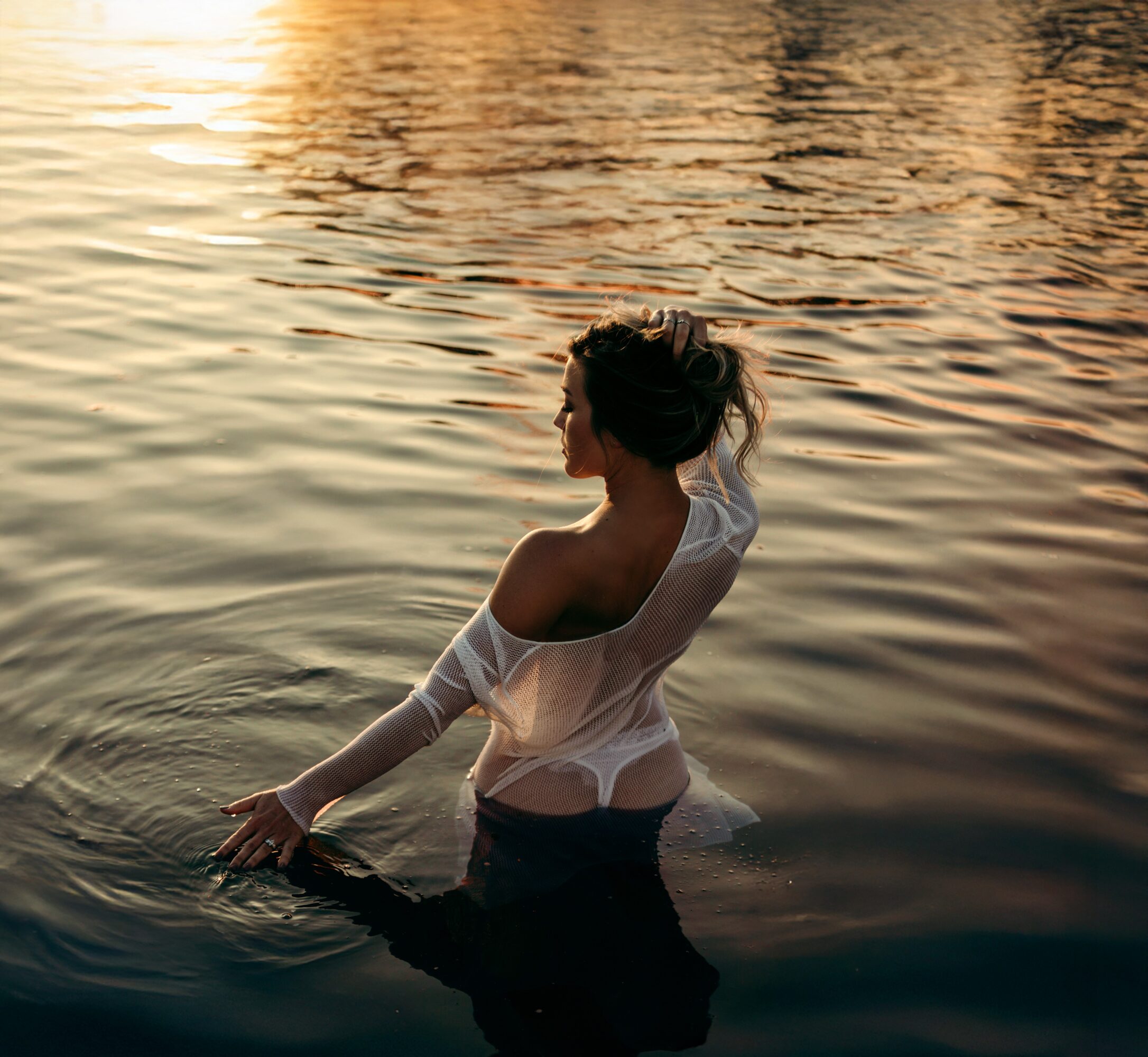 Woman standing in water during sunset, wearing a white off-shoulder top, with her hair tied up, facing sideways.