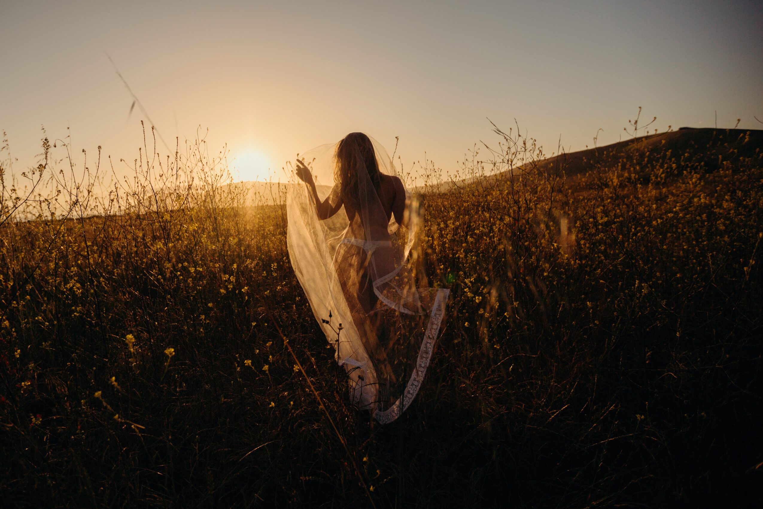 Person walking through a grassy field at sunset, holding a flowing fabric, with a hill in the background.