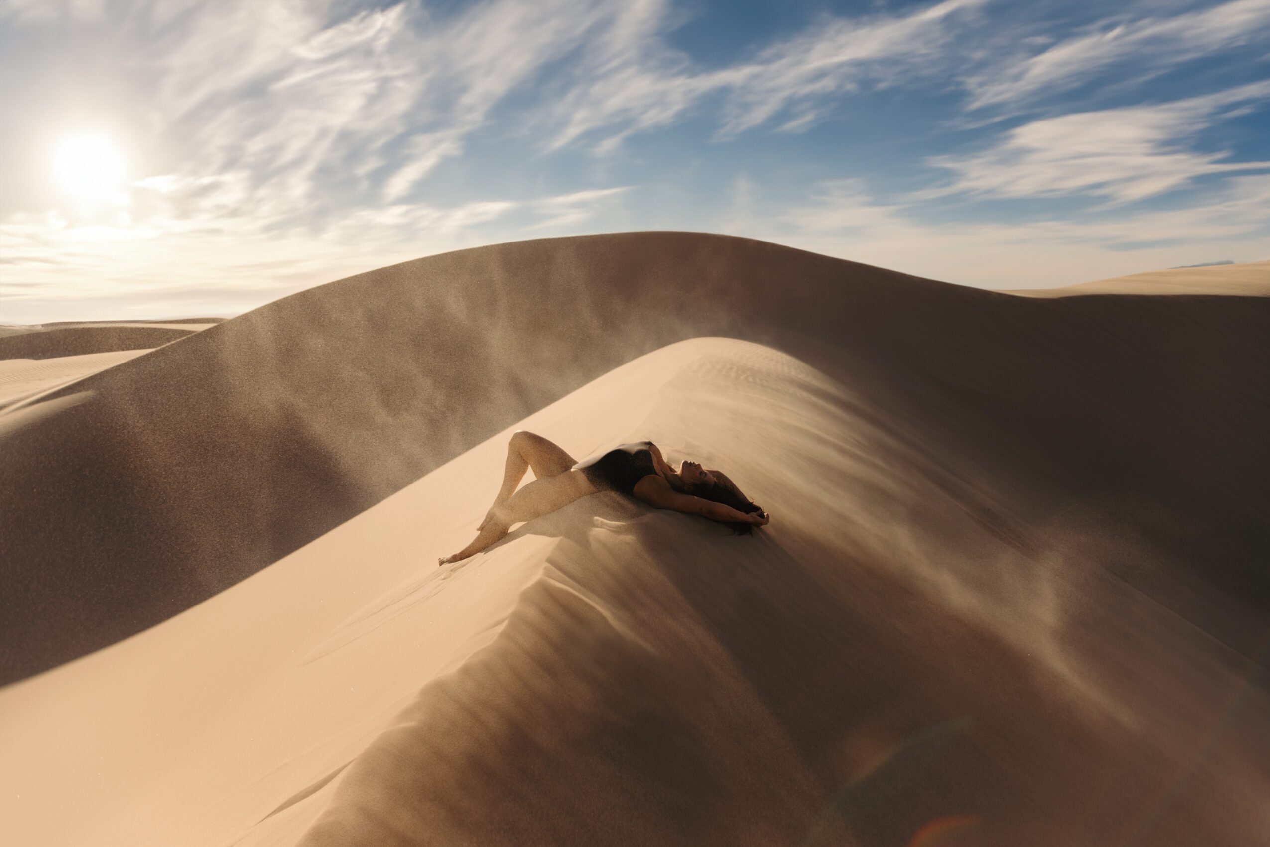 Person lying on sand dune with wind-blown sand, desert landscape, blue sky with clouds, sun shining.