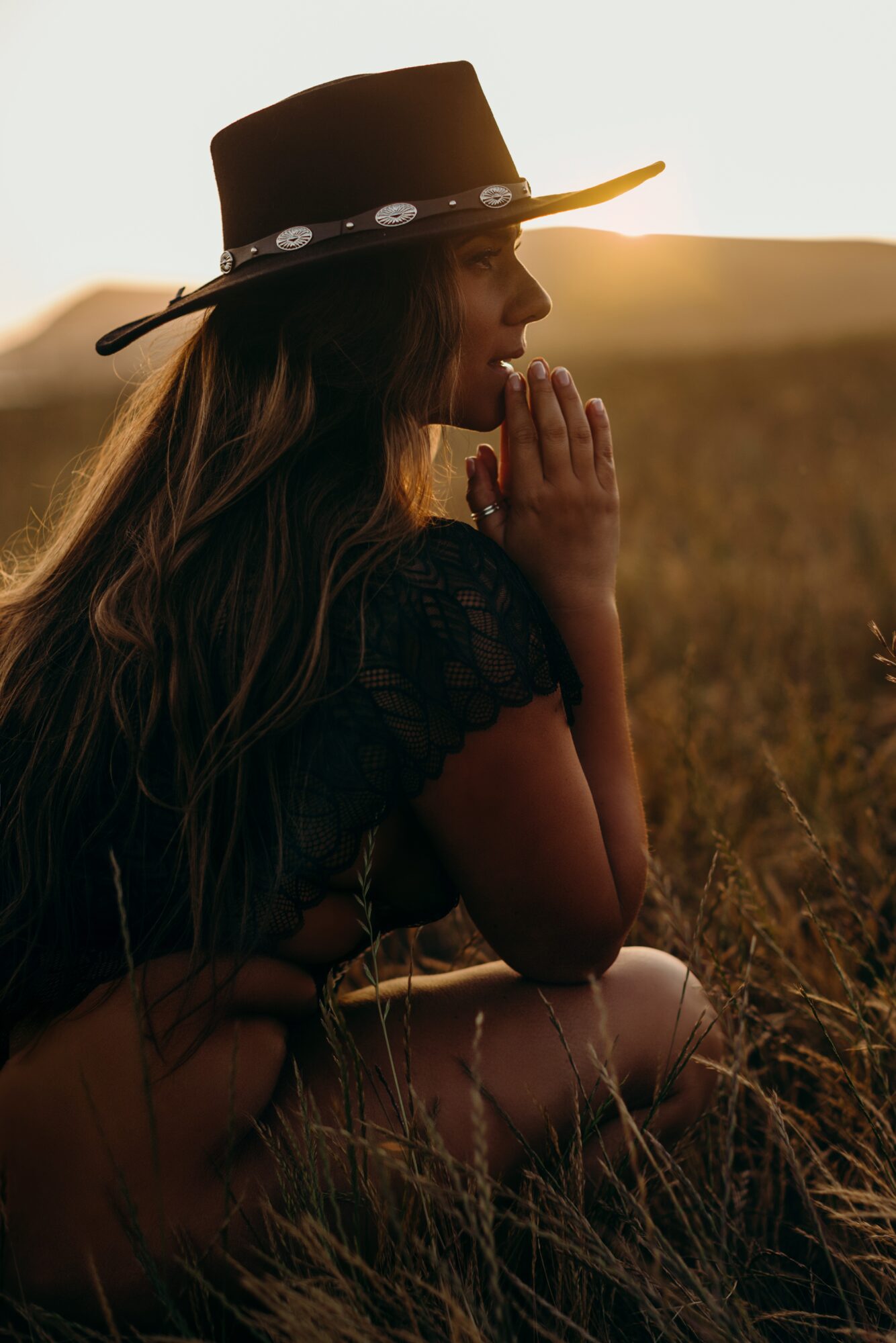 Woman wearing a hat, sitting in a field during sunset, with hands clasped near her face, looking contemplative.