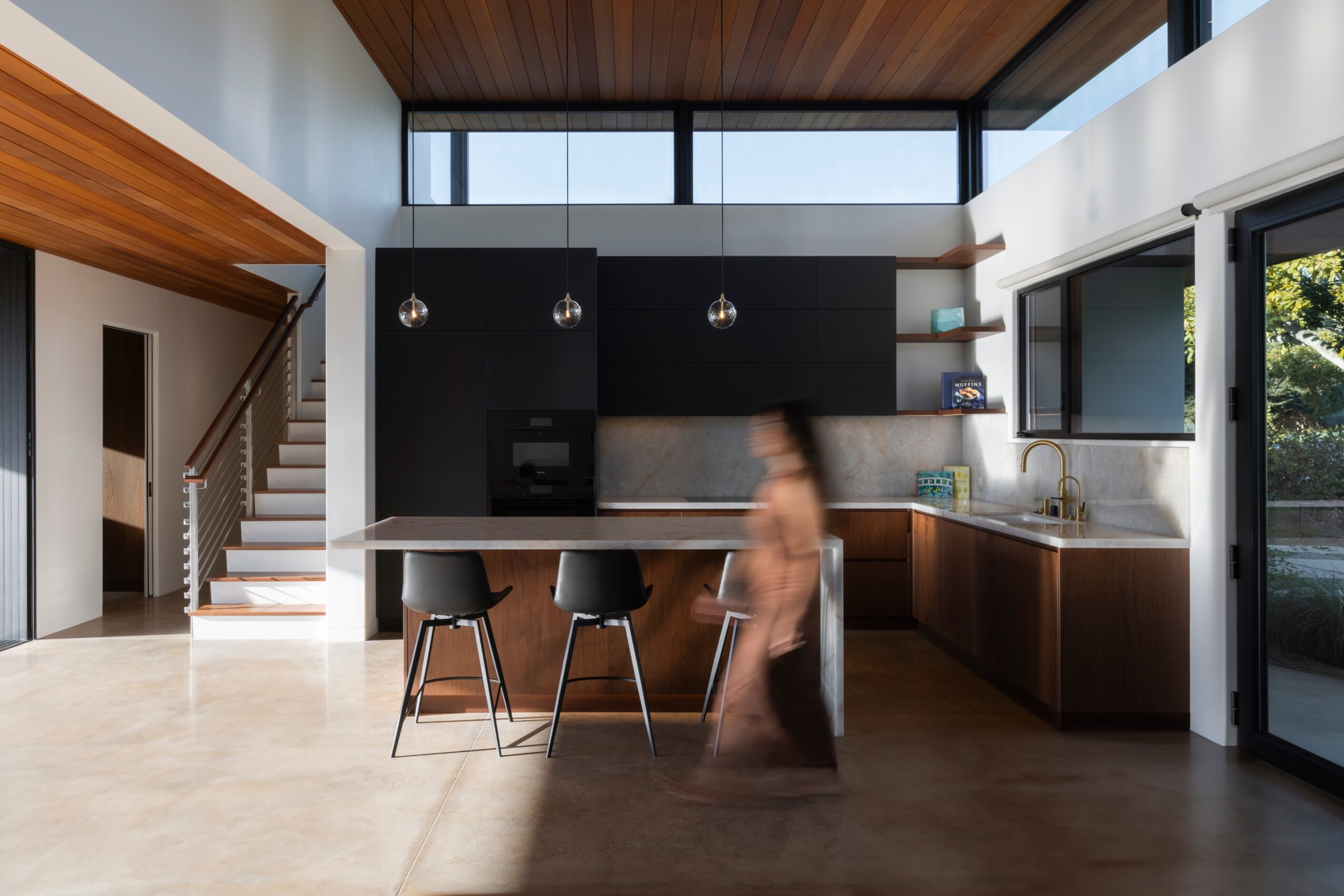 Modern kitchen with black cabinets, a large window, and a person walking past the island, with a staircase visible on the left.