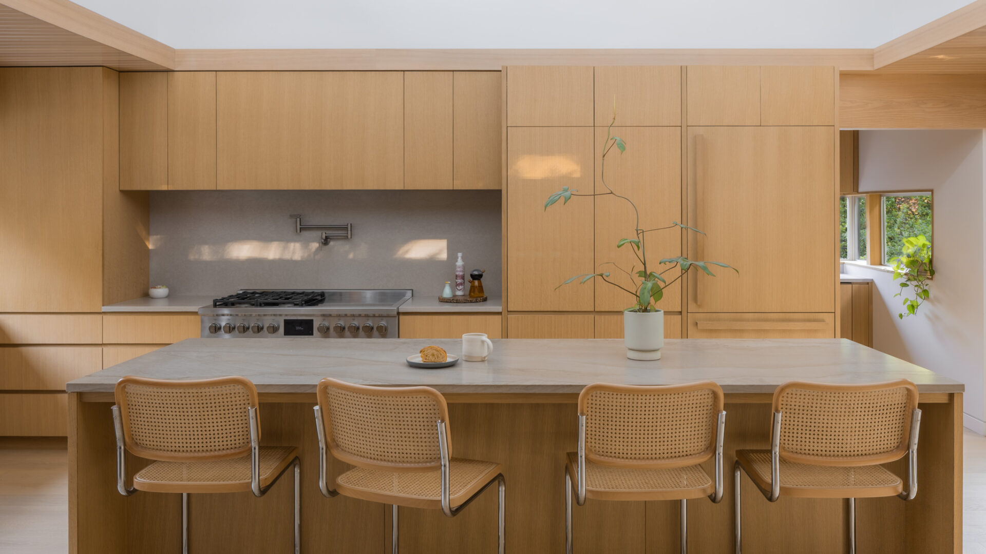 Kitchen with wooden cabinets, a white countertop, and four chairs at a breakfast bar, with a potted plant and a view outside.