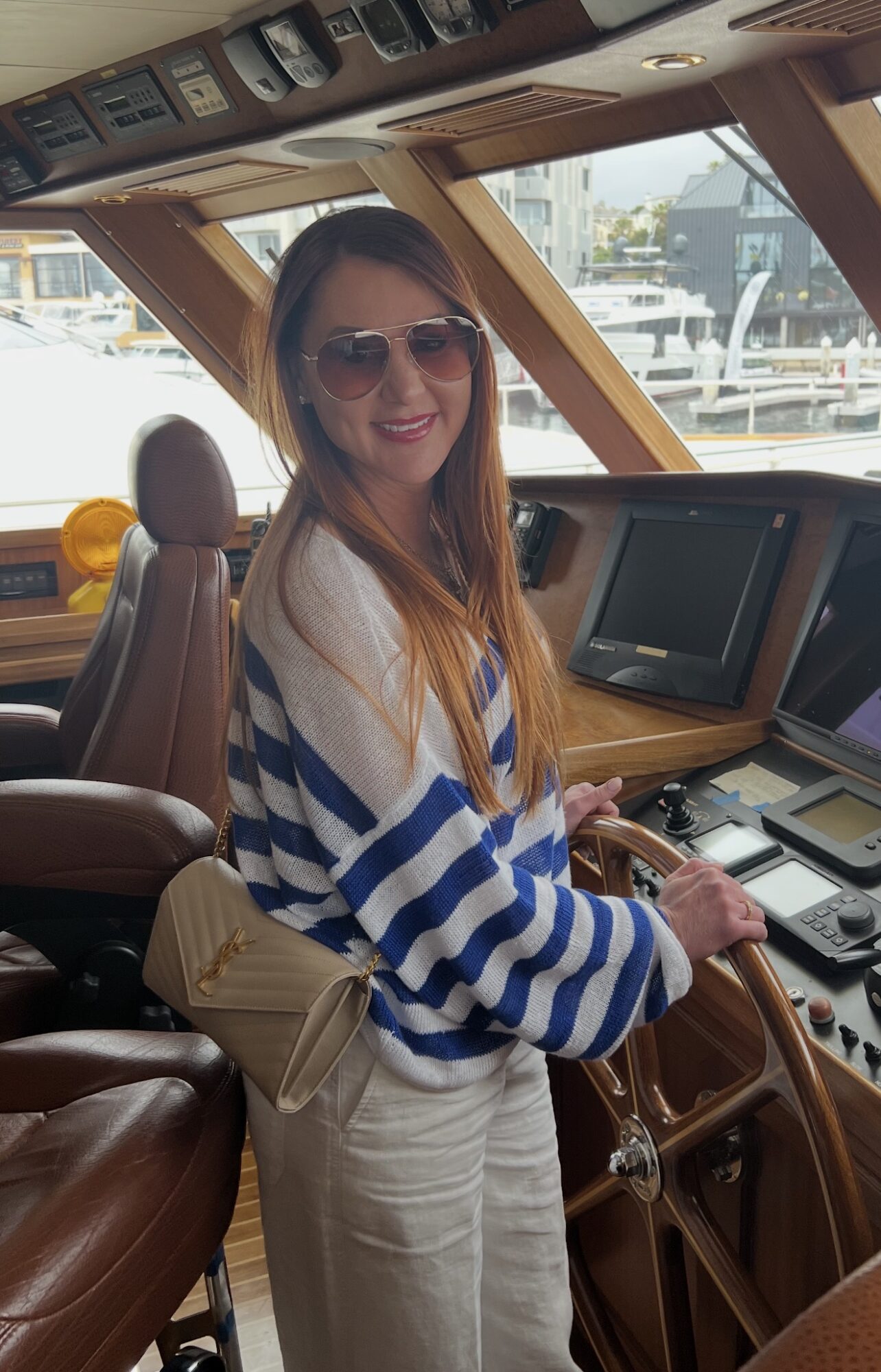 Woman with sunglasses smiling inside a boat's control room, wearing a striped sweater and light-colored pants.