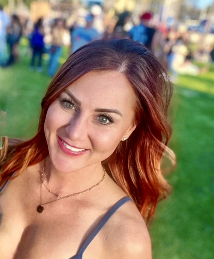 Woman with red hair smiling outdoors, background with people and green grass