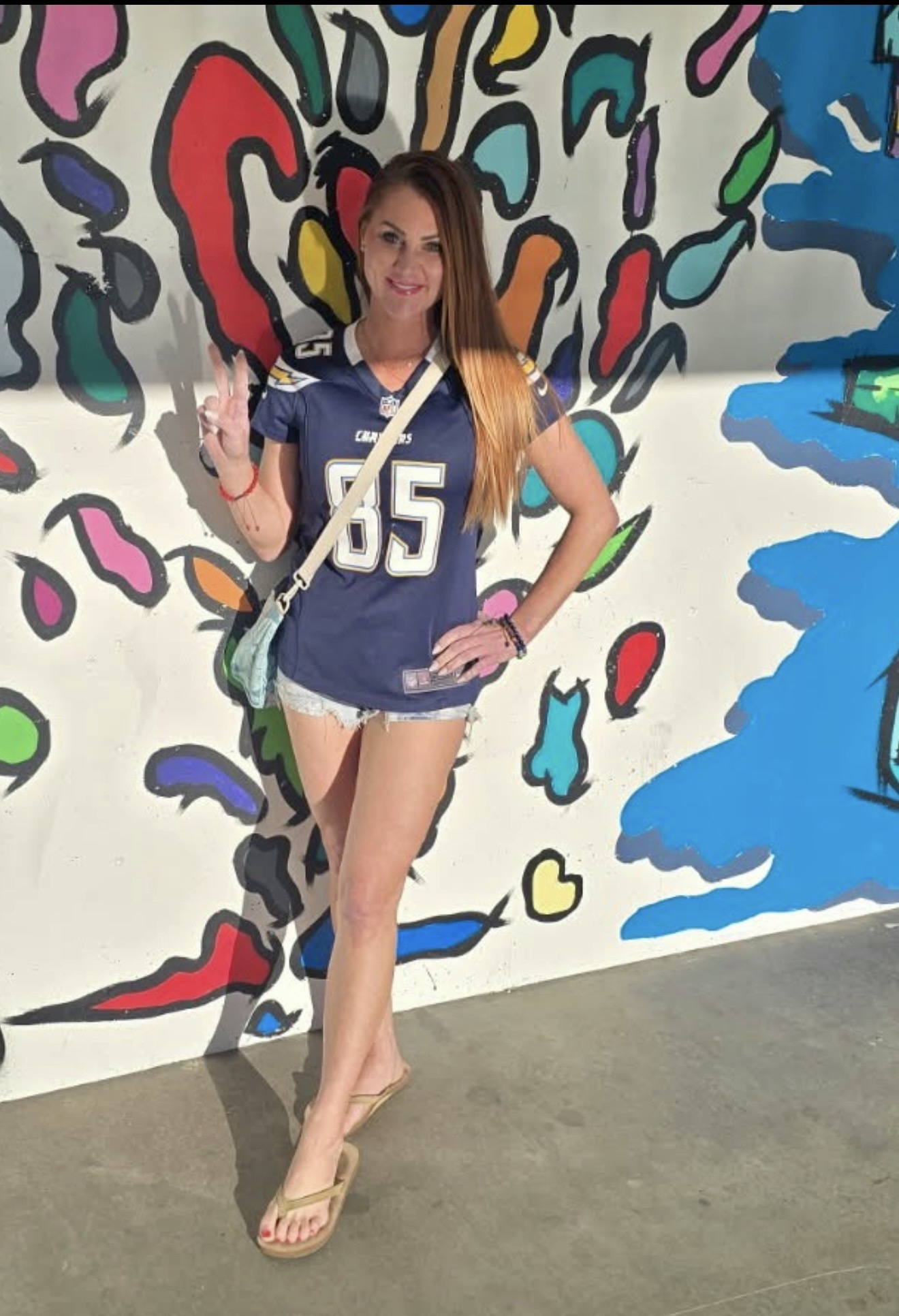 Young woman with long hair standing in front of a colorful abstract mural, smiling, wearing a blue sports jersey and shorts.