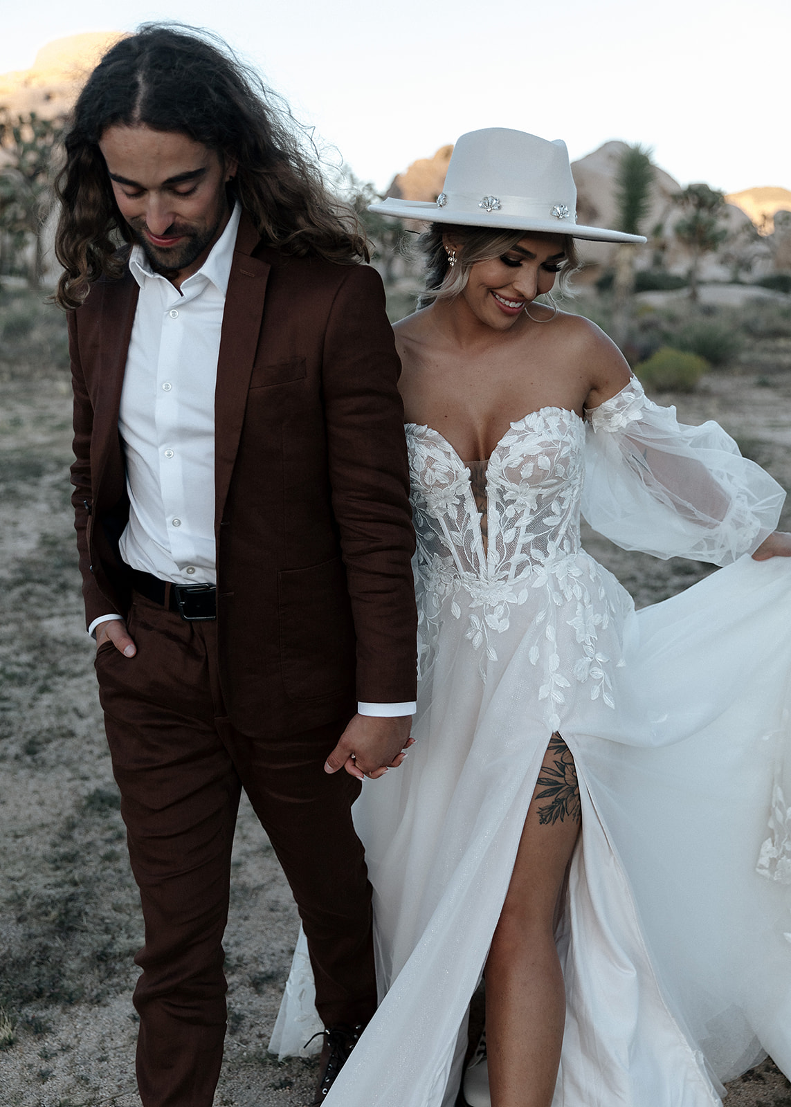 A couple holding hands outdoors, the woman in a white dress and wide-brimmed hat, the man in a brown suit, both smiling.