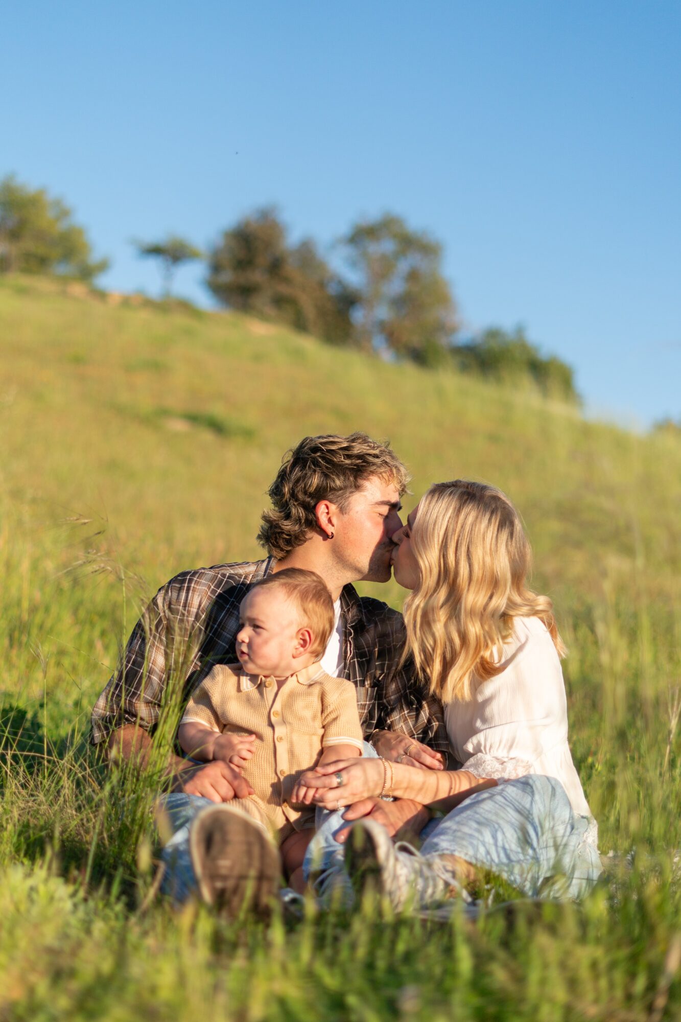 Family sitting on grass in a field, with a man, woman, and baby, under a clear blue sky.