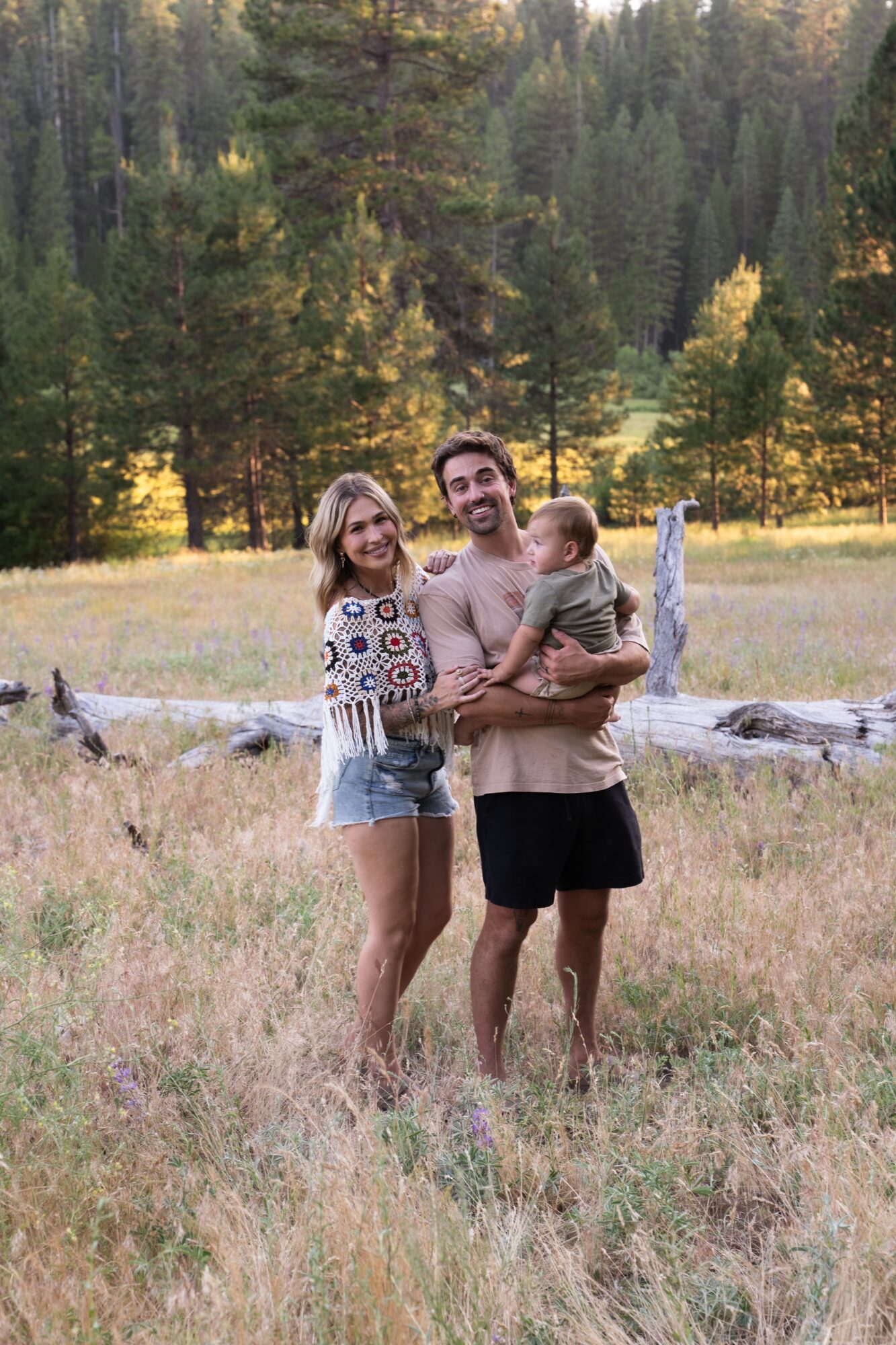Family of three standing outdoors in a grassy field with trees in the background, smiling at the camera.
