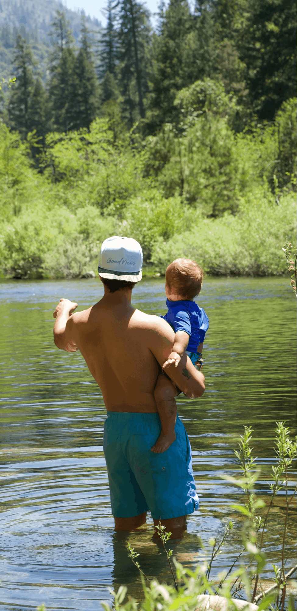 Person holding a child in a river with trees and greenery in the background.