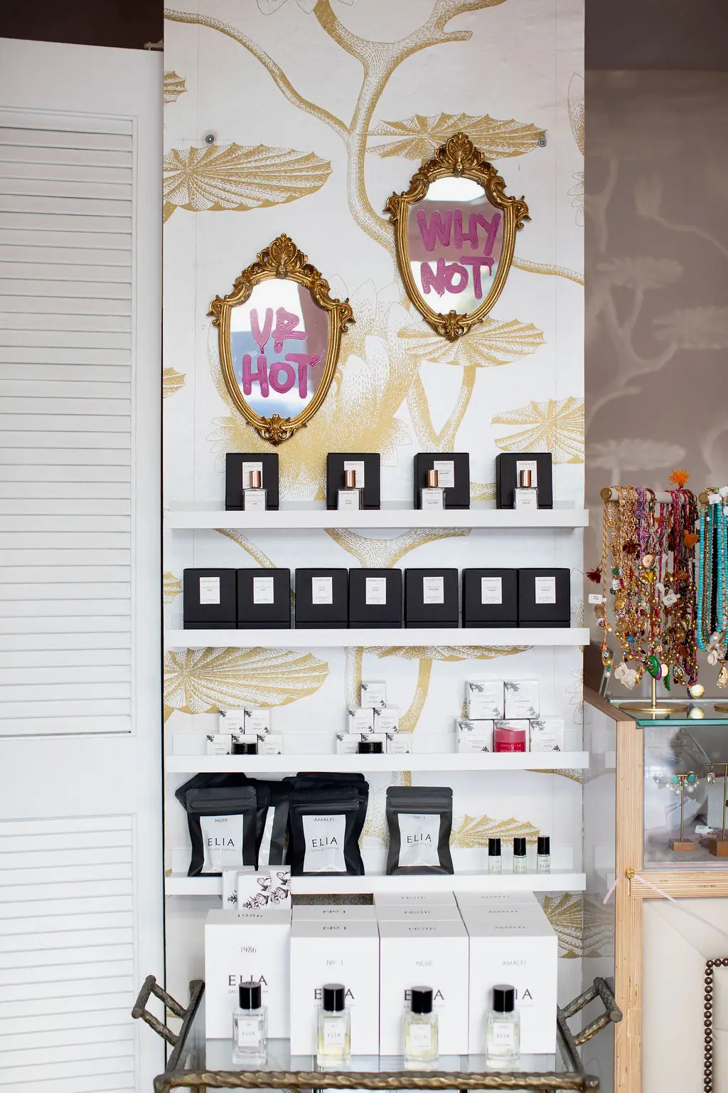 Shelves with beauty products and decorative mirrors against a patterned wall, with a white louvered door on the left. Word signs read 'WHY NOT'.