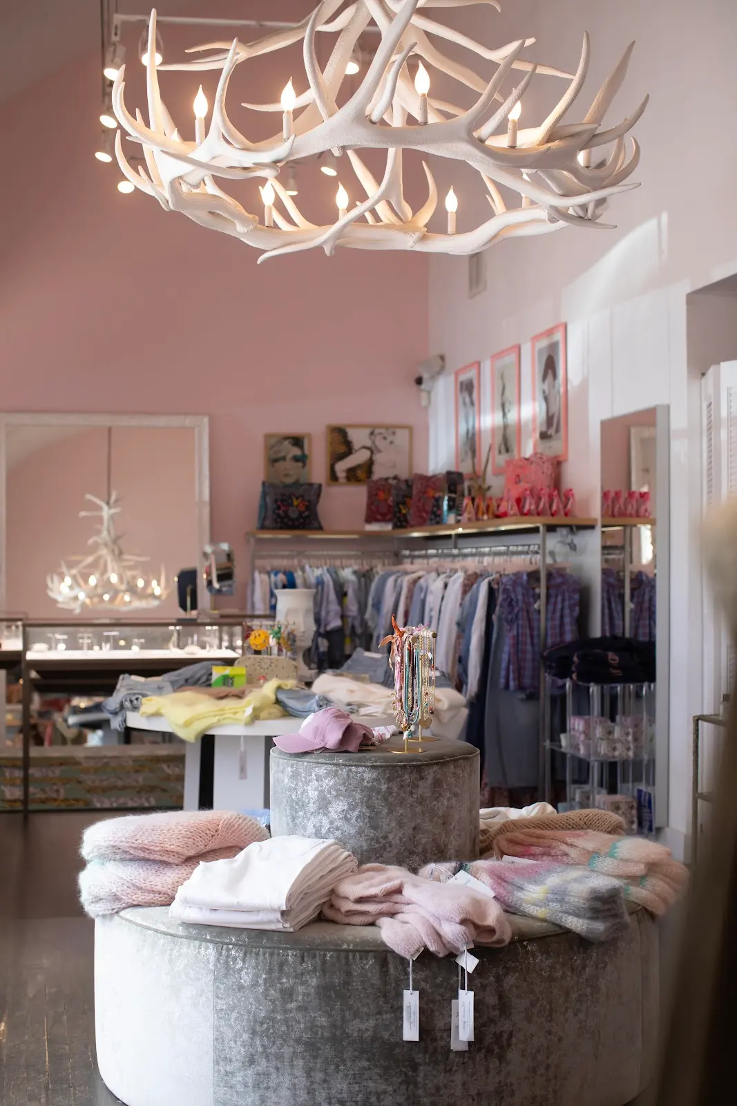 Clothing store interior with a chandelier, display table, and racks of clothes, illuminated by natural light from windows.