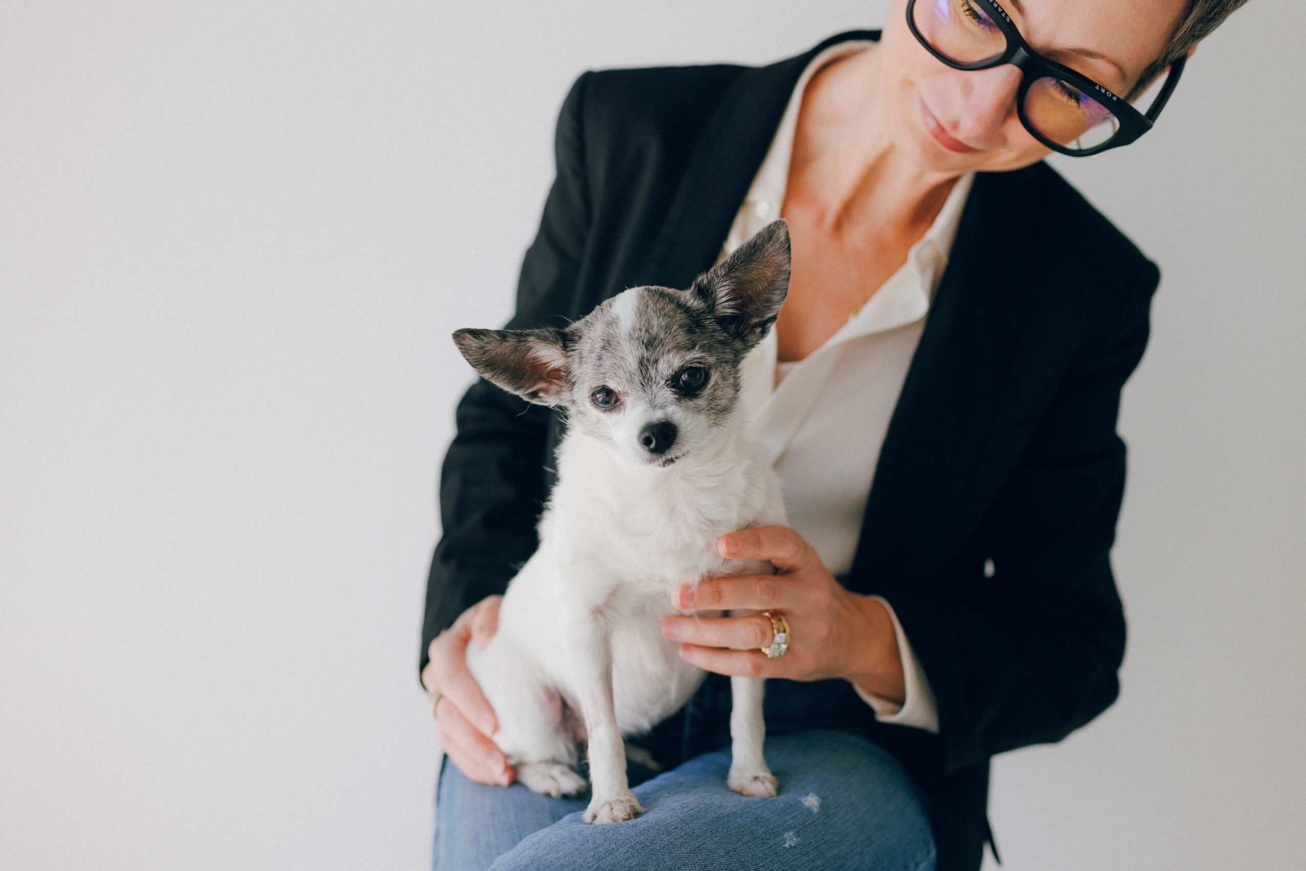 Person holding a small dog, wearing glasses and a dark blazer, against a plain background.