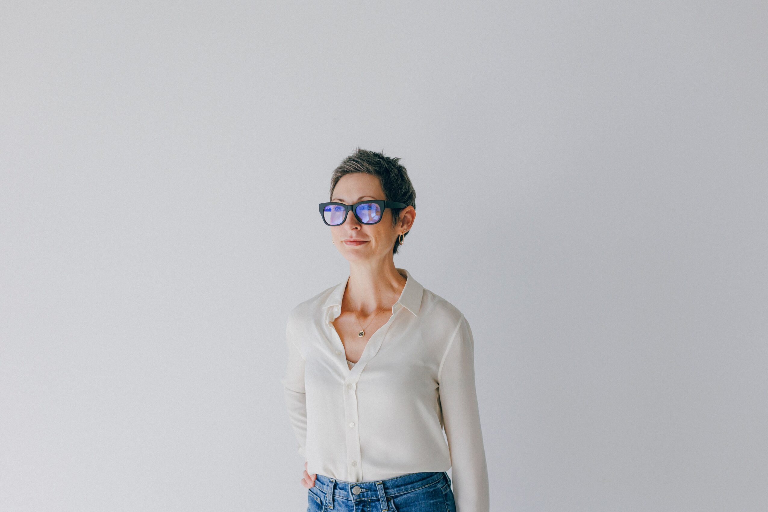 Woman with short dark hair wearing glasses and a white blouse, standing against a plain light gray background.