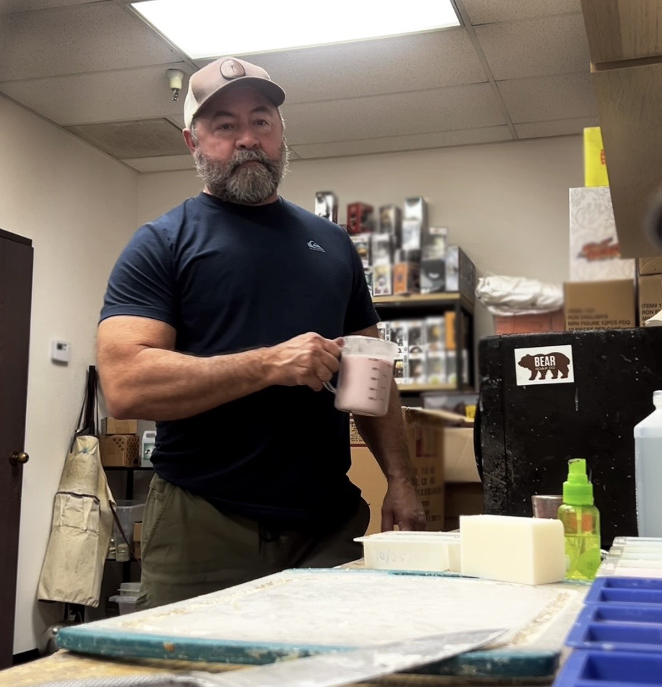 Man with beard and cap holding a mug in a room with shelves and boxes behind him.
