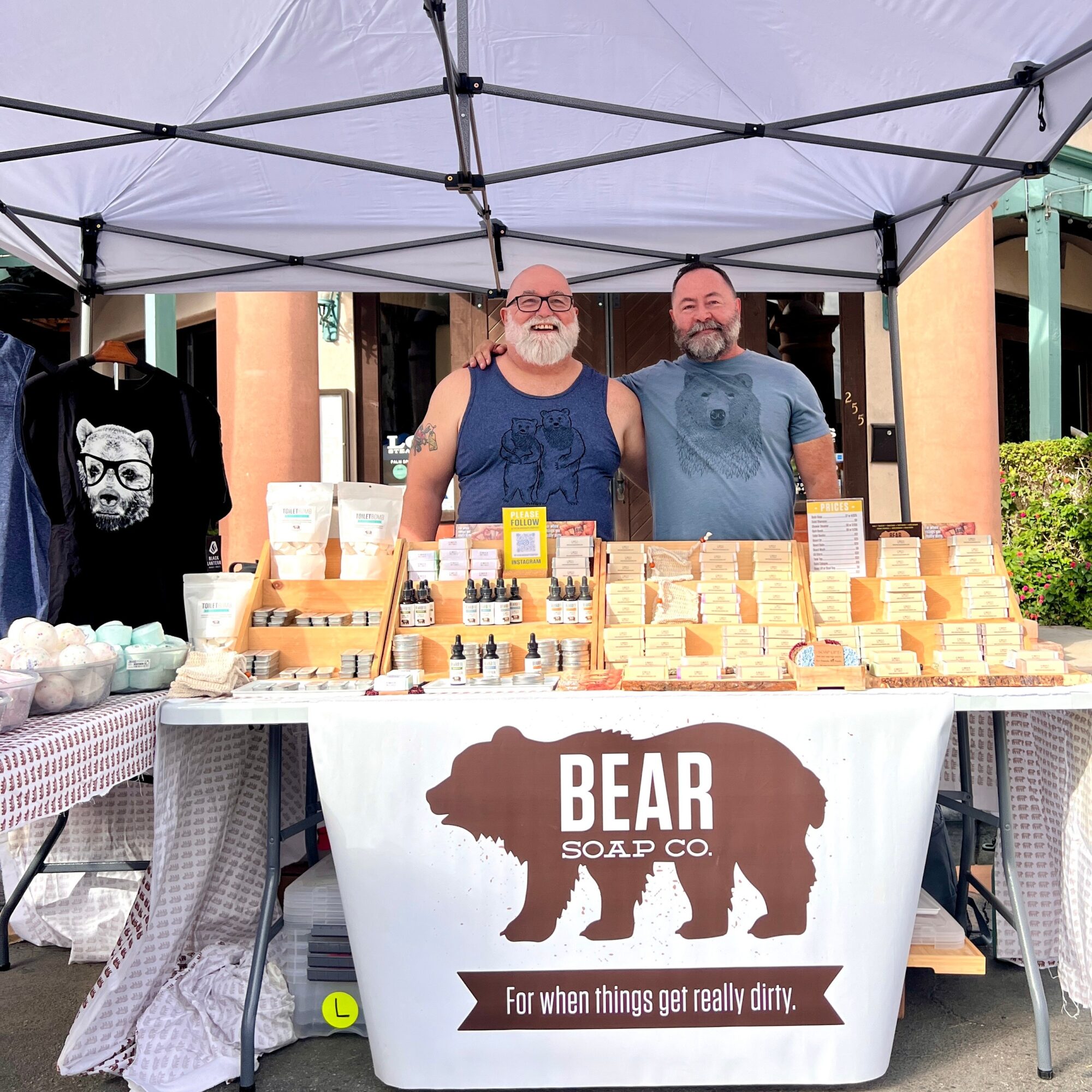 Two men stand behind a soap booth with a bear logo, displaying soap products outdoors under a canopy.
