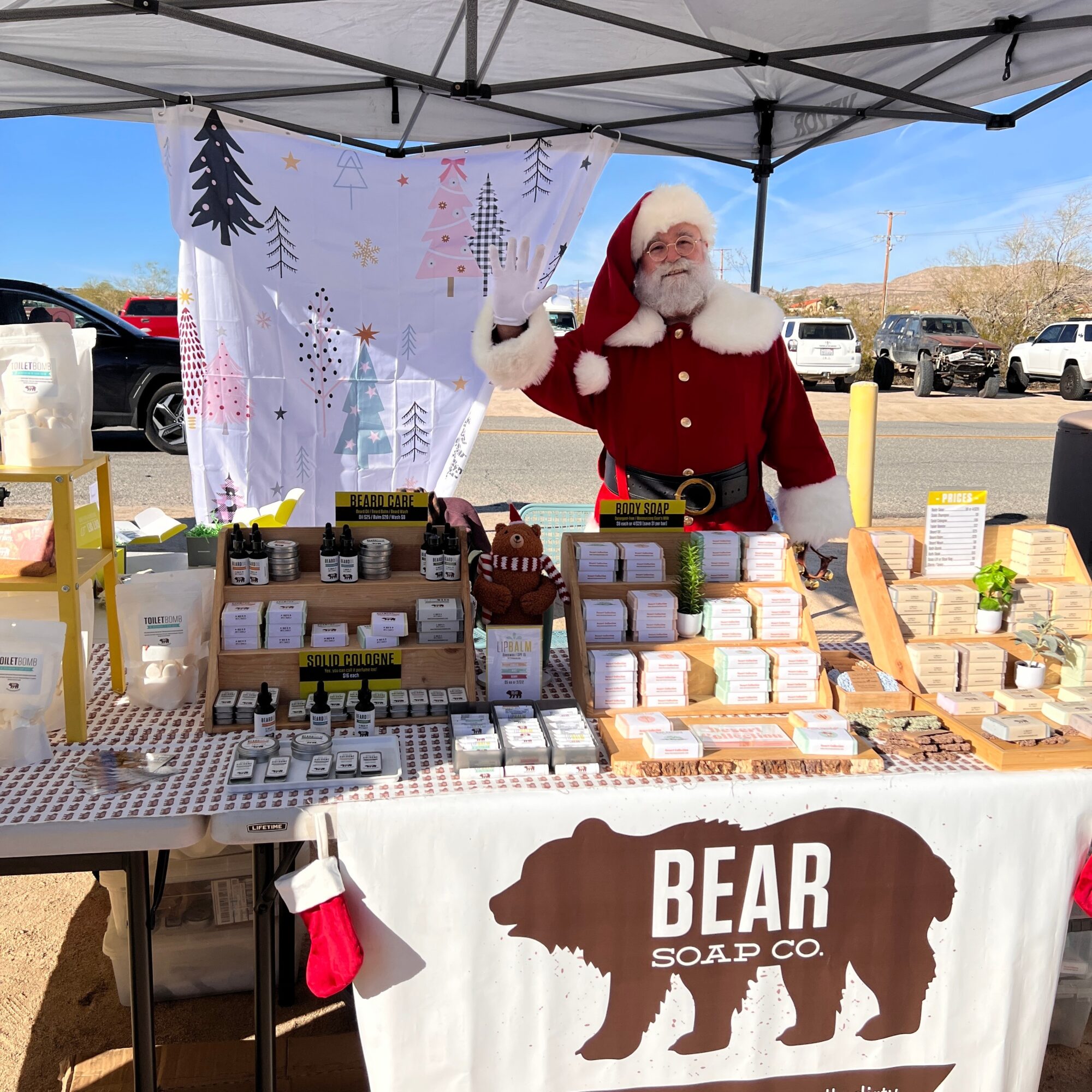 Santa Claus standing behind a table with soap products at an outdoor market stall, under a canopy with a winter-themed backdrop.