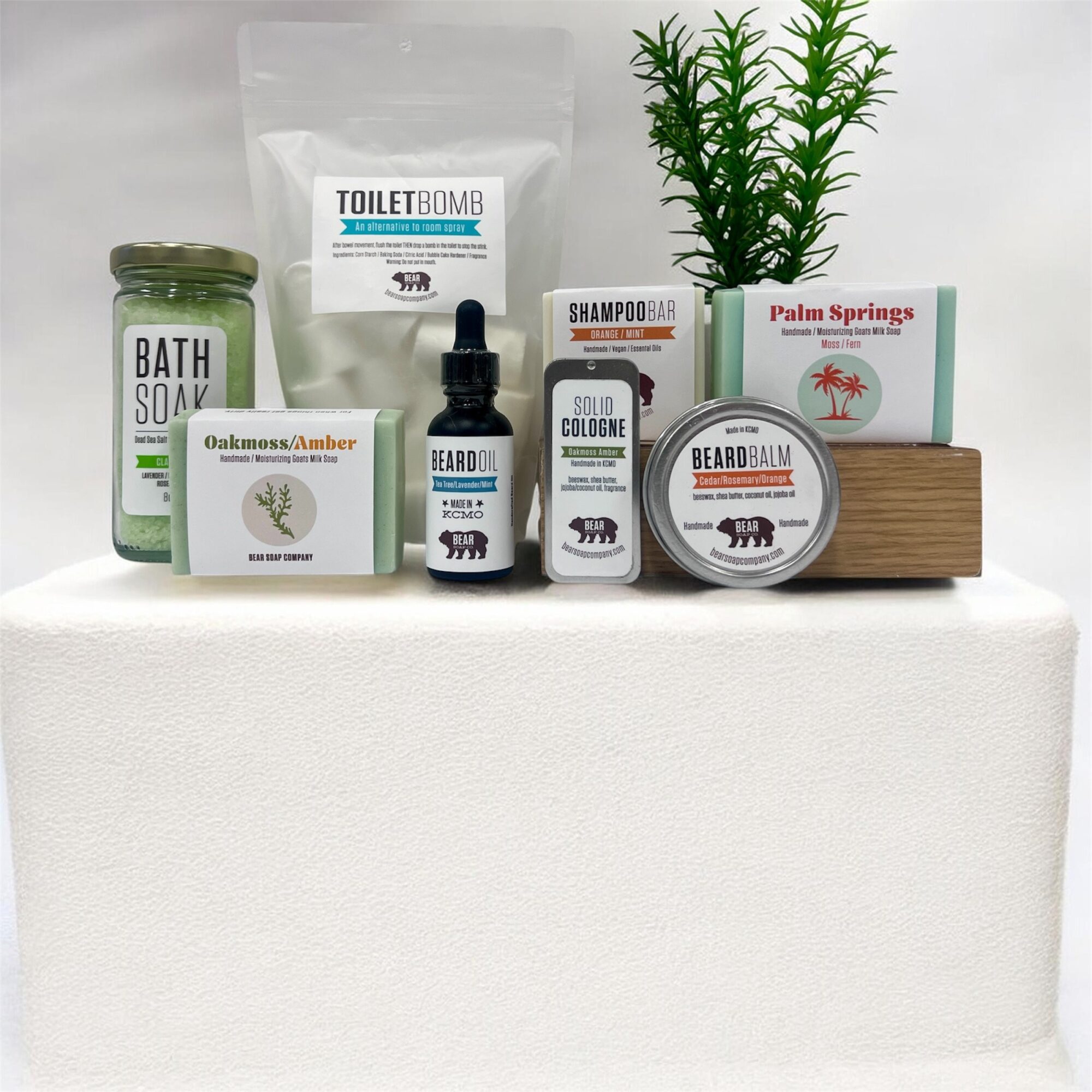 Collection of personal care products, a green plant, and a wooden box on a white surface, against a white background.