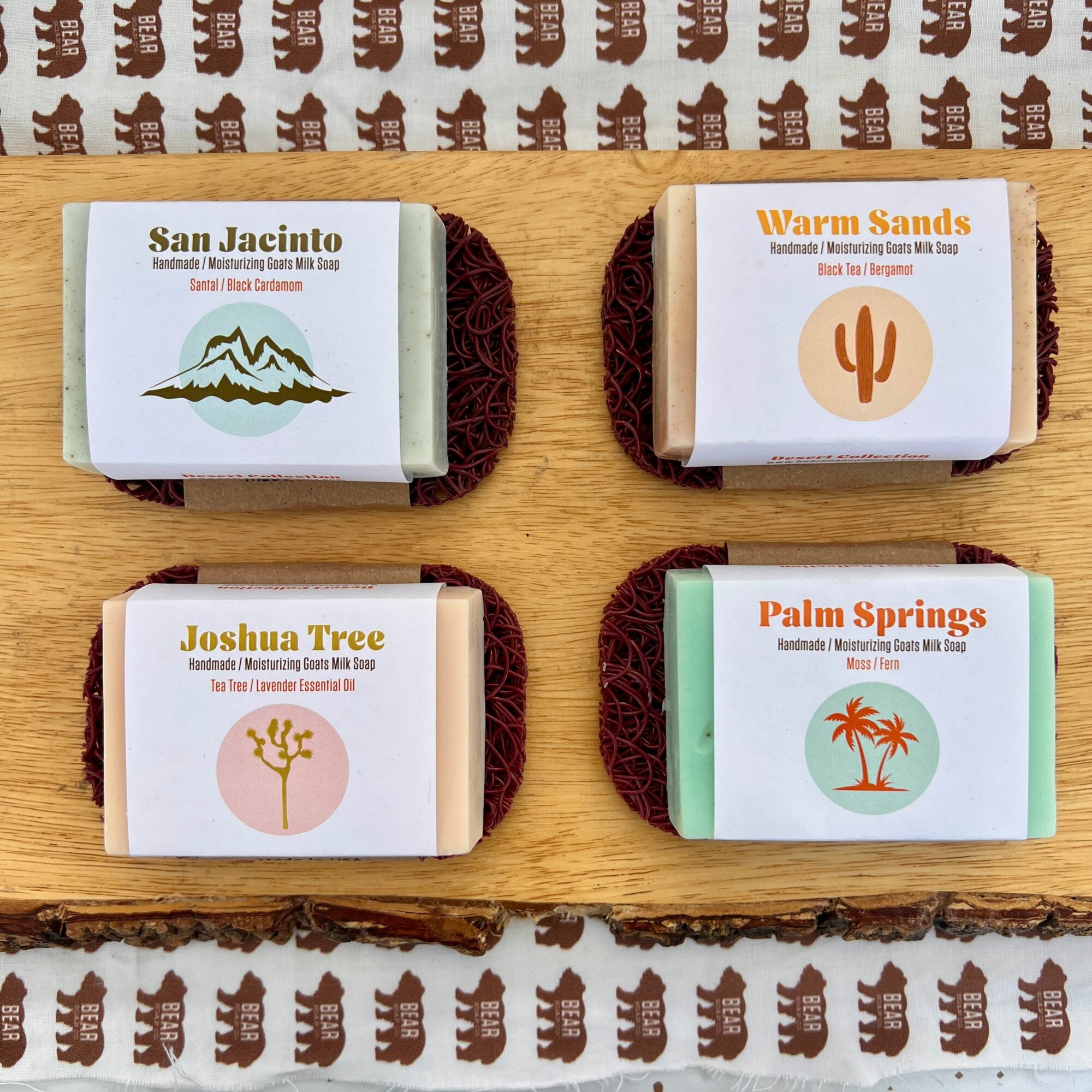 Four soap bars with desert and mountain themes on a wooden surface, each wrapped with a label and a textured base.