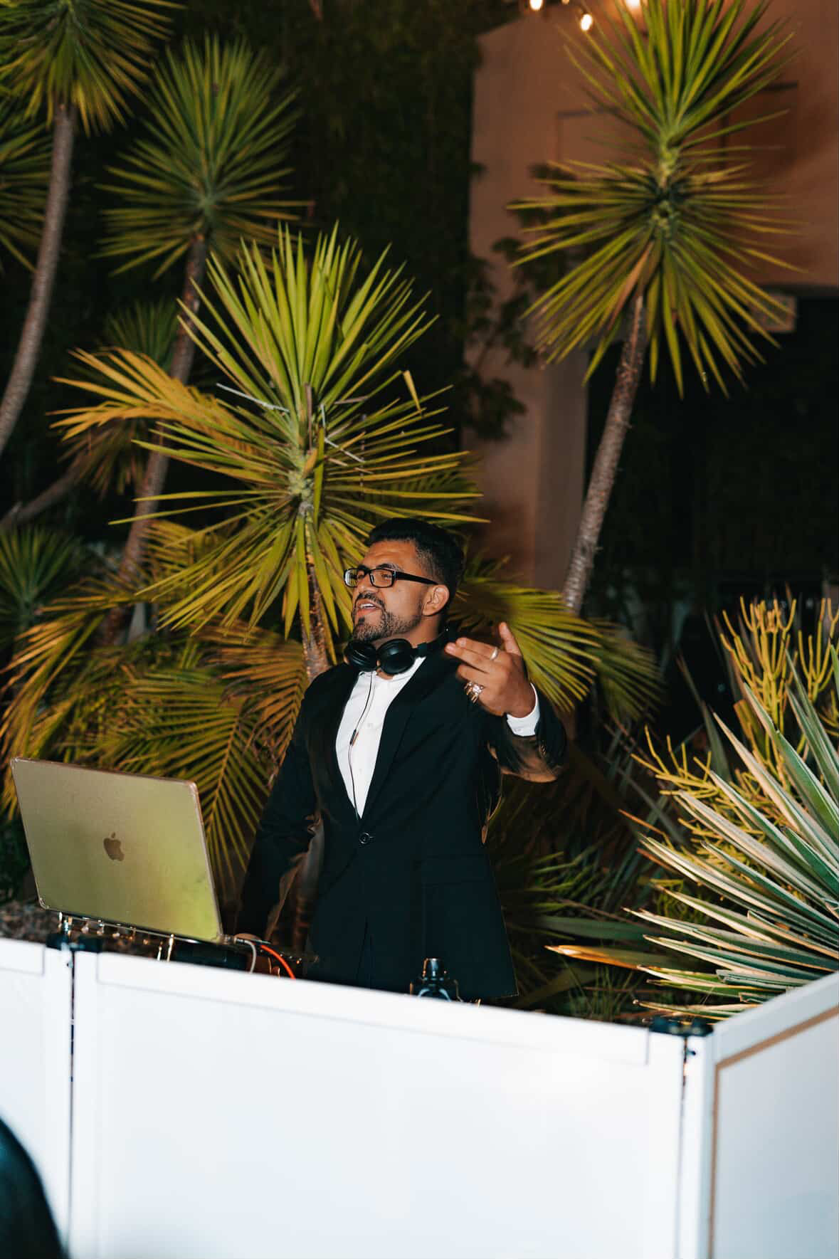 Man in tuxedo with glasses standing behind a DJ booth outdoors at night, surrounded by palm trees.