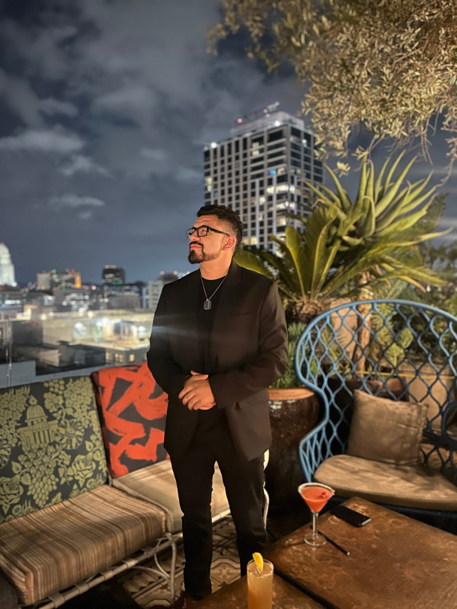 Man in black suit with glasses standing on rooftop terrace at night, city skyline and tall building in background, plants, and seating area with drinks.