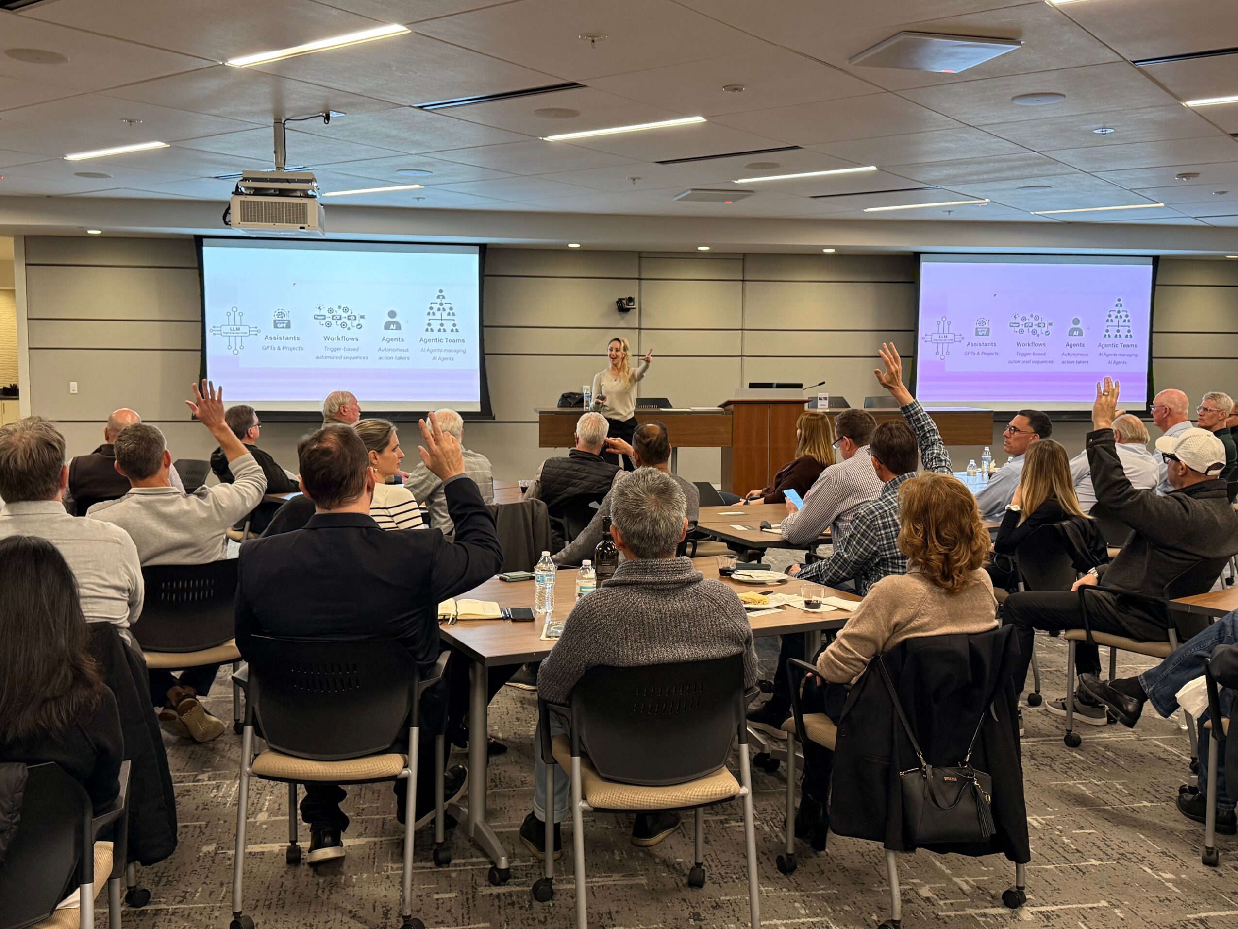Conference room with a speaker presenting to an audience, two large screens display presentation slides, attendees seated at tables, some raising hands.