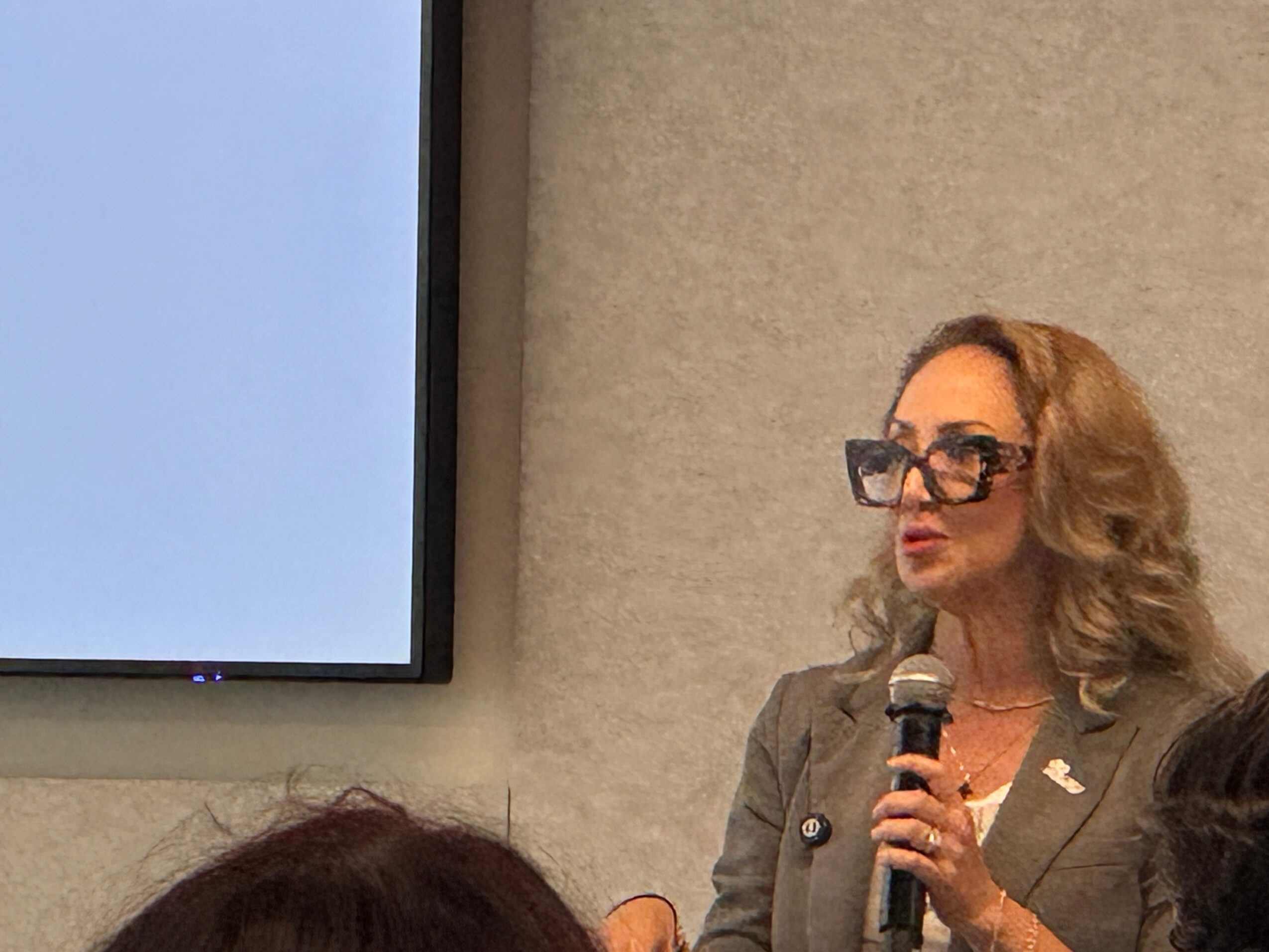 Woman with glasses holding a microphone, speaking in front of a screen and a gray wall.