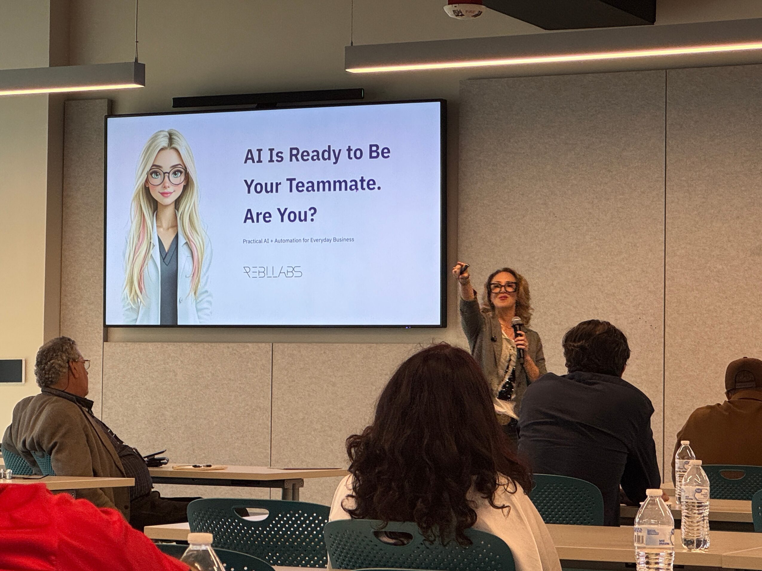 Woman giving a presentation in a conference room with a large screen displaying a slide about AI and a woman with glasses. Four people are seated, listening. Word count: 20.