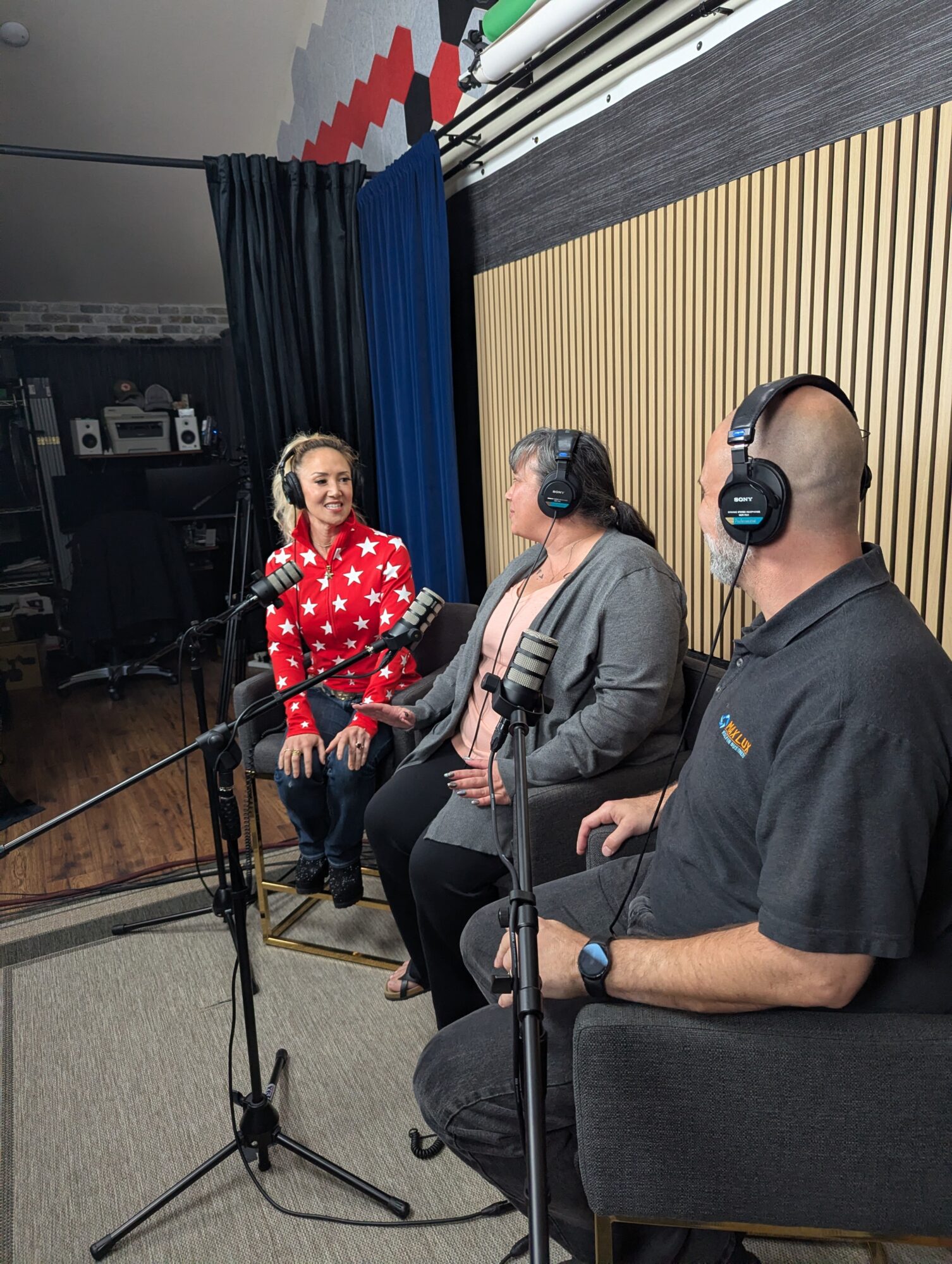 Three people sit in a recording studio with microphones, headphones, and a wooden wall background.