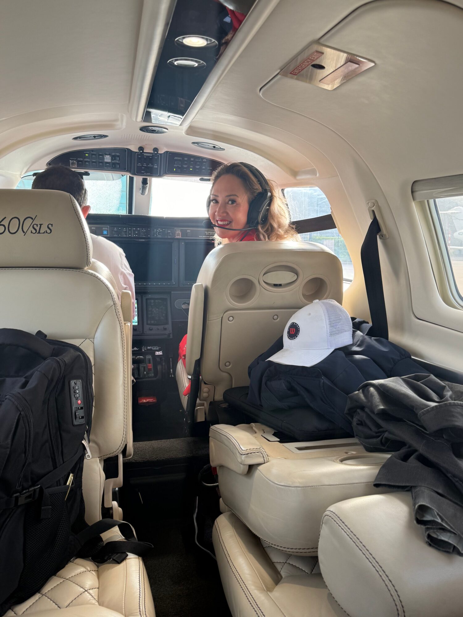 Person sitting in airplane seat with window view, surrounded by bags and personal items, facing forward.
