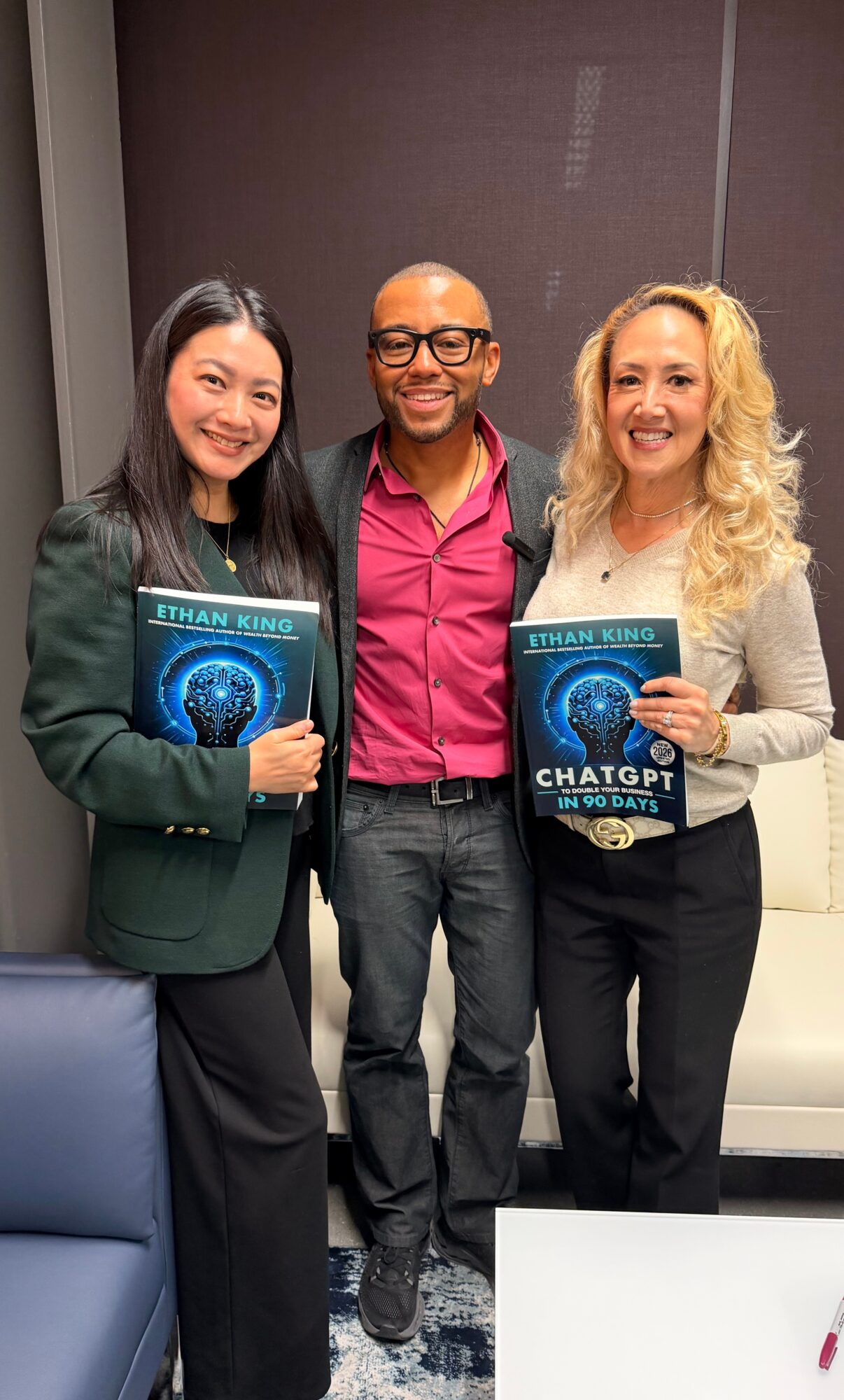 Three people standing indoors, two women holding books, smiling, with a man in the middle, in front of a gray wall.