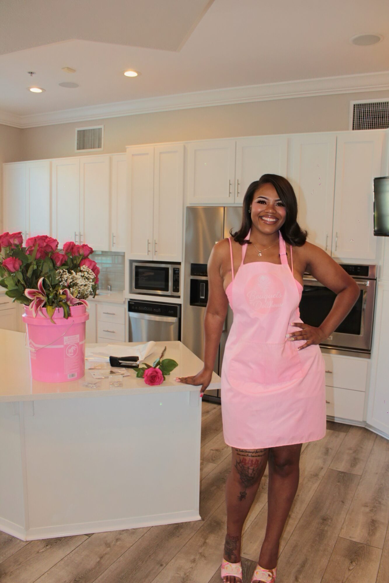 Woman in pink apron standing in modern kitchen with white cabinets and flowers on counter.
