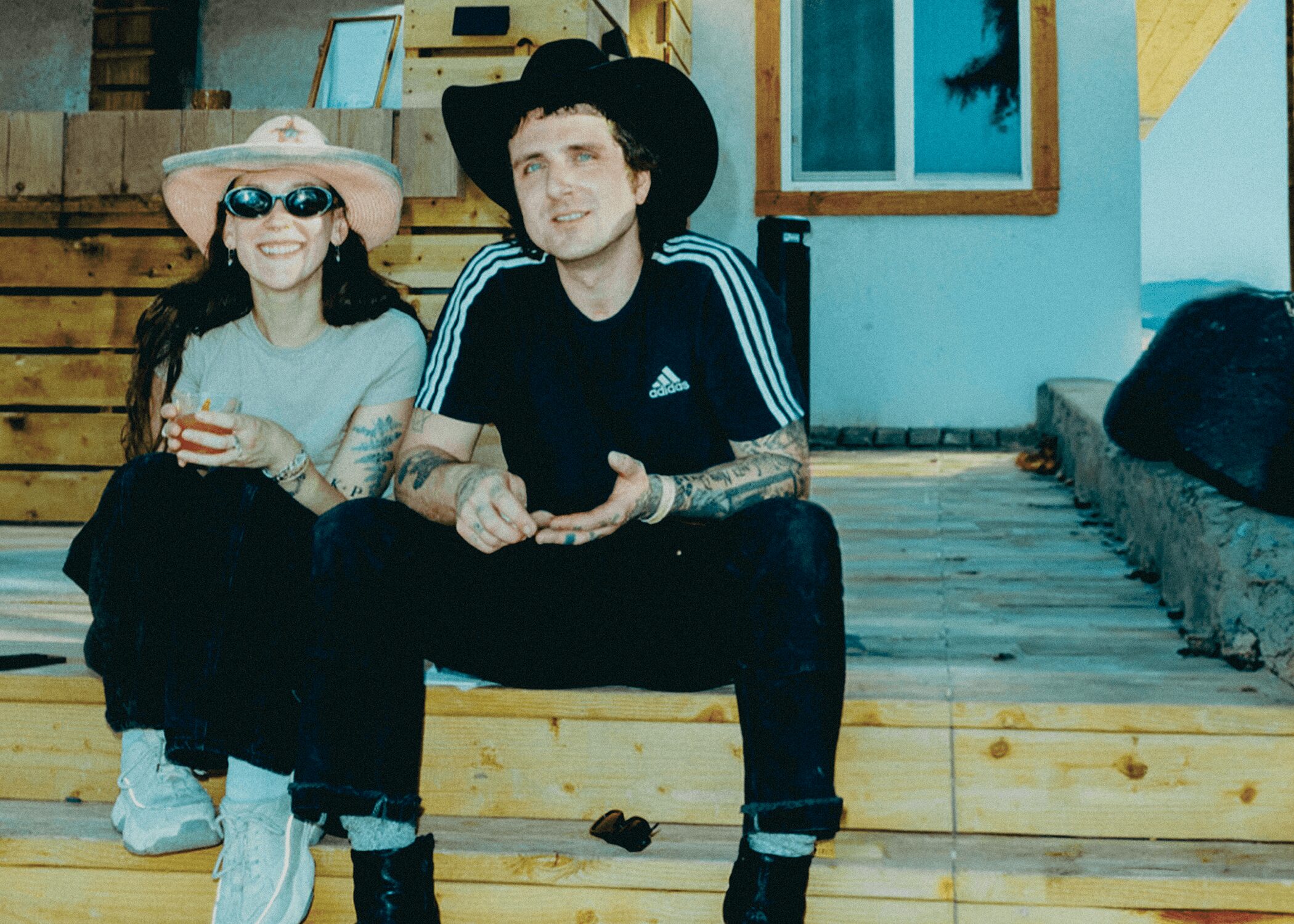 Two people sitting on wooden steps outside a house, one wearing a hat and sunglasses, the other in a cowboy hat.