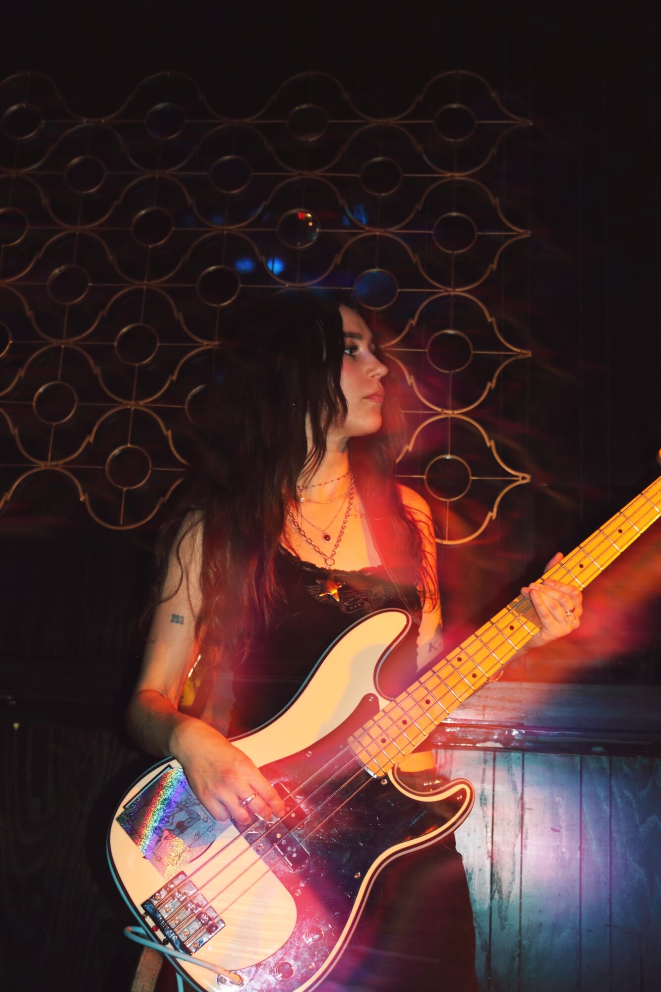 Woman with long dark hair playing an electric guitar in a dimly lit room with decorative wall art behind her.
