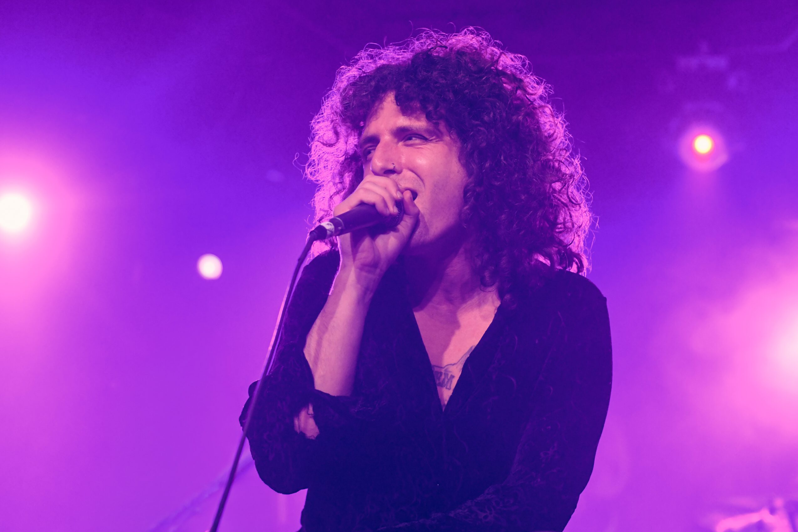 Person with curly hair singing into a microphone on stage with purple and pink lighting, eyes closed, expressive performance.