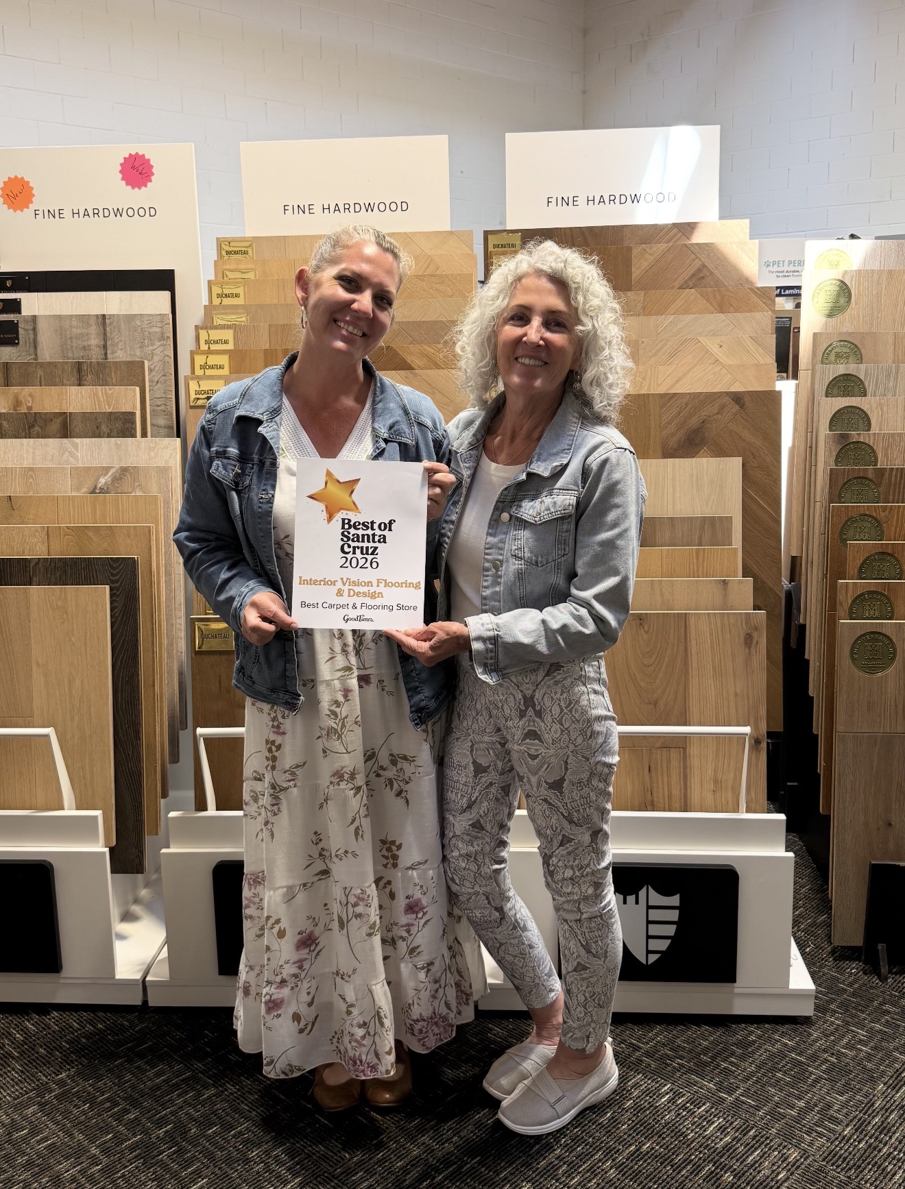 Two women stand in front of wood sample displays, smiling and holding a certificate. One wears a floral dress, the other a patterned suit.
