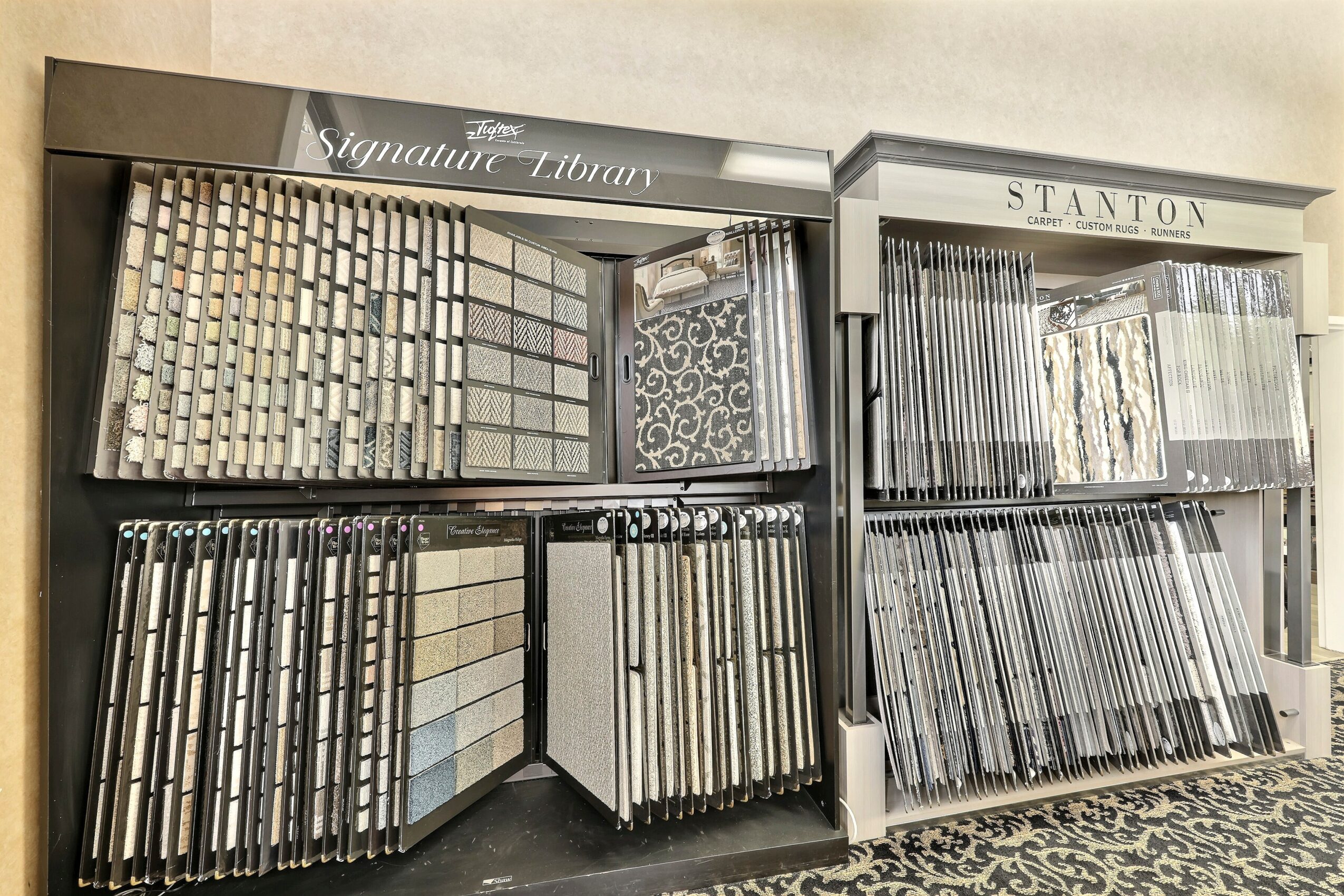 Display of various tile samples arranged on shelves in a store, with signage above, and a patterned carpet below.