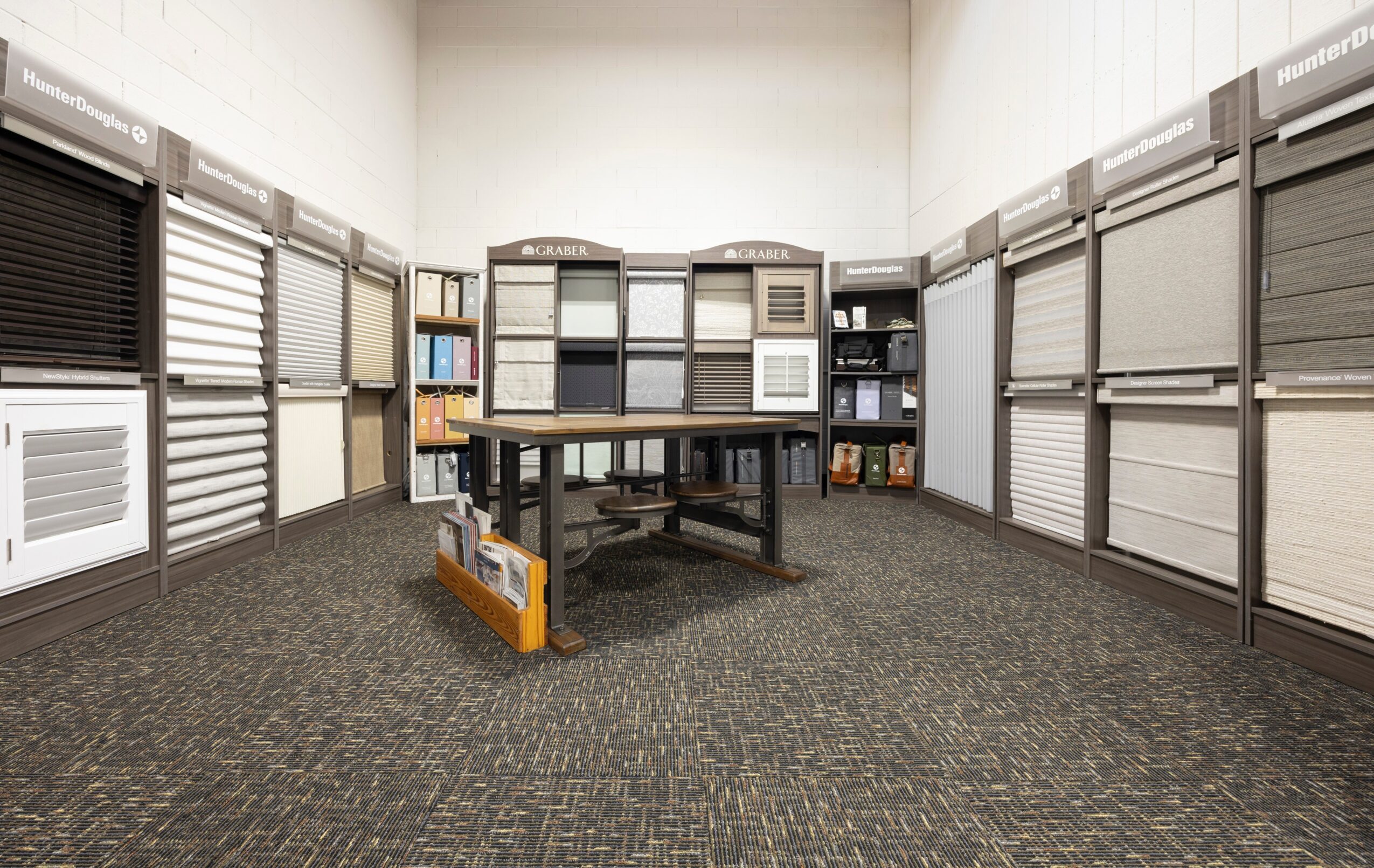 Display room with window blinds and window coverings on walls, table with samples, and carpeted floor.
