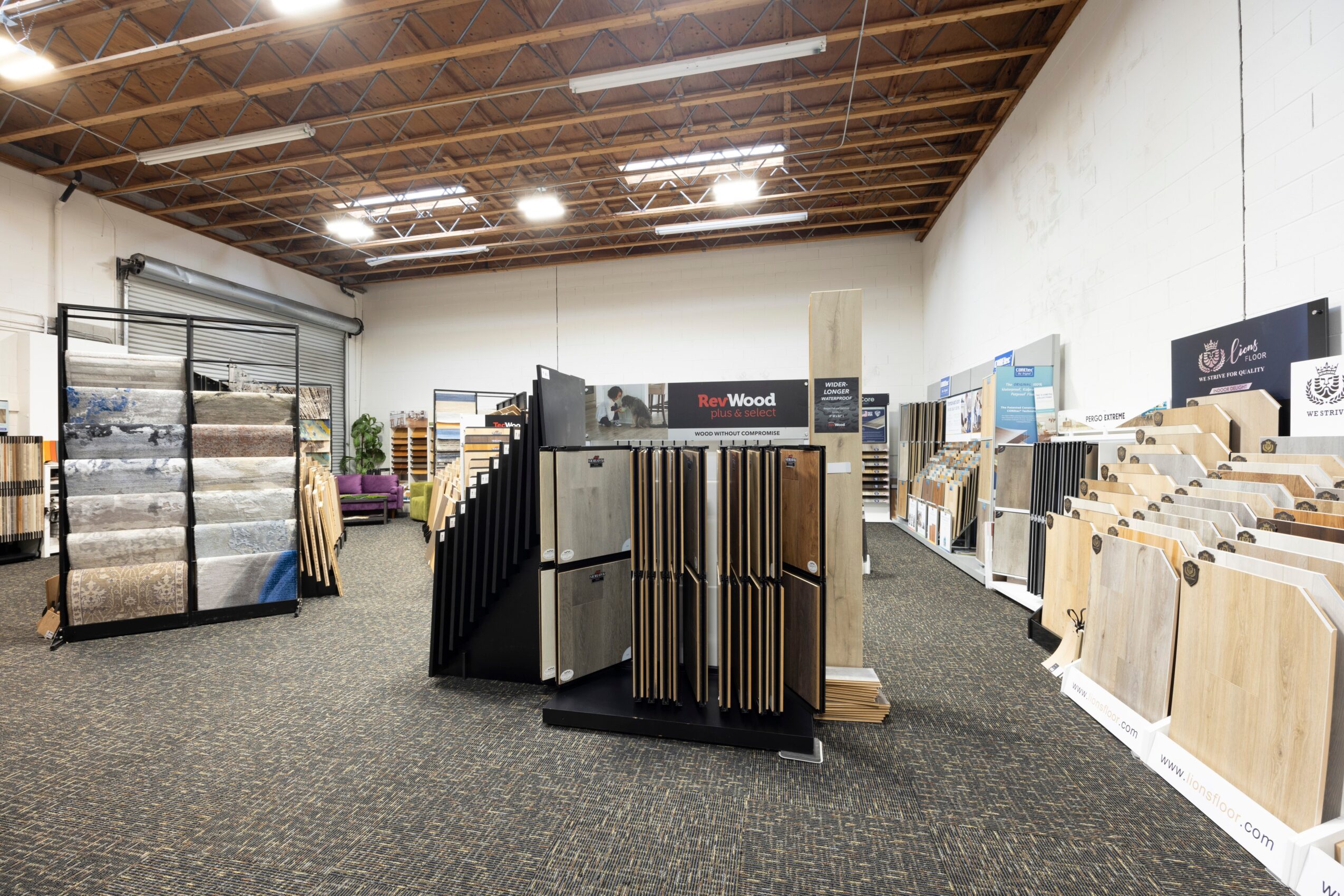 Interior of a store with flooring samples, display racks, and a white wall, ceiling with exposed wood beams.
