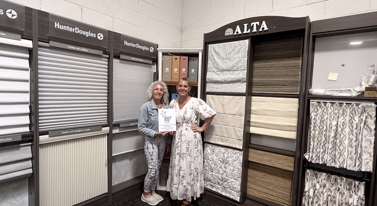 Two women standing in a showroom with display samples of window blinds and curtains, smiling at the camera.