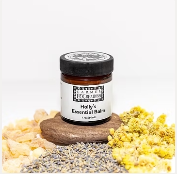 Small jar labeled Holly's Essential Balm surrounded by yellow flowers, lavender, and a brown stone on a white background.