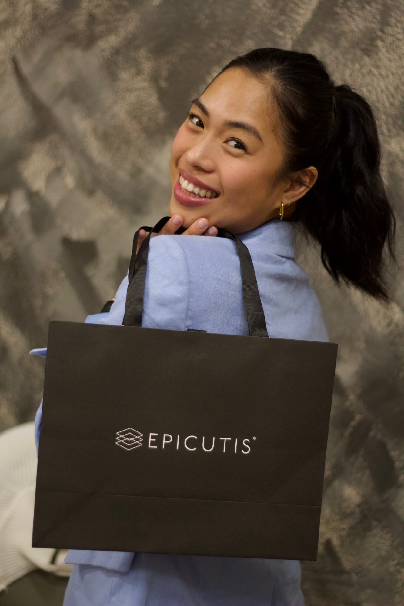 Smiling woman holding a black Epicitis shopping bag, wearing a light blue top, with a textured background.