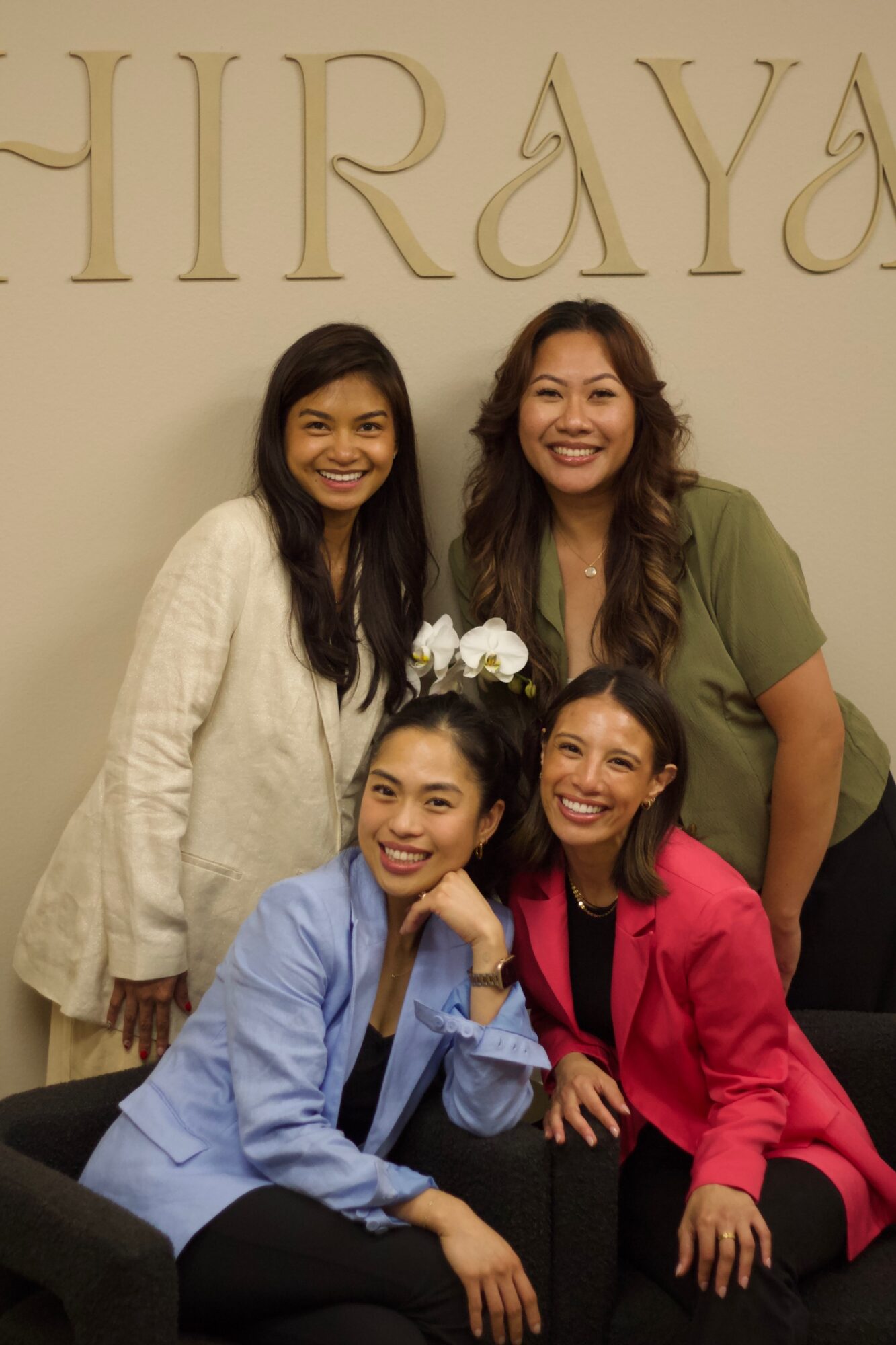 Four women smiling, sitting and standing in front of a wall with large gold letters, posing for a photo.