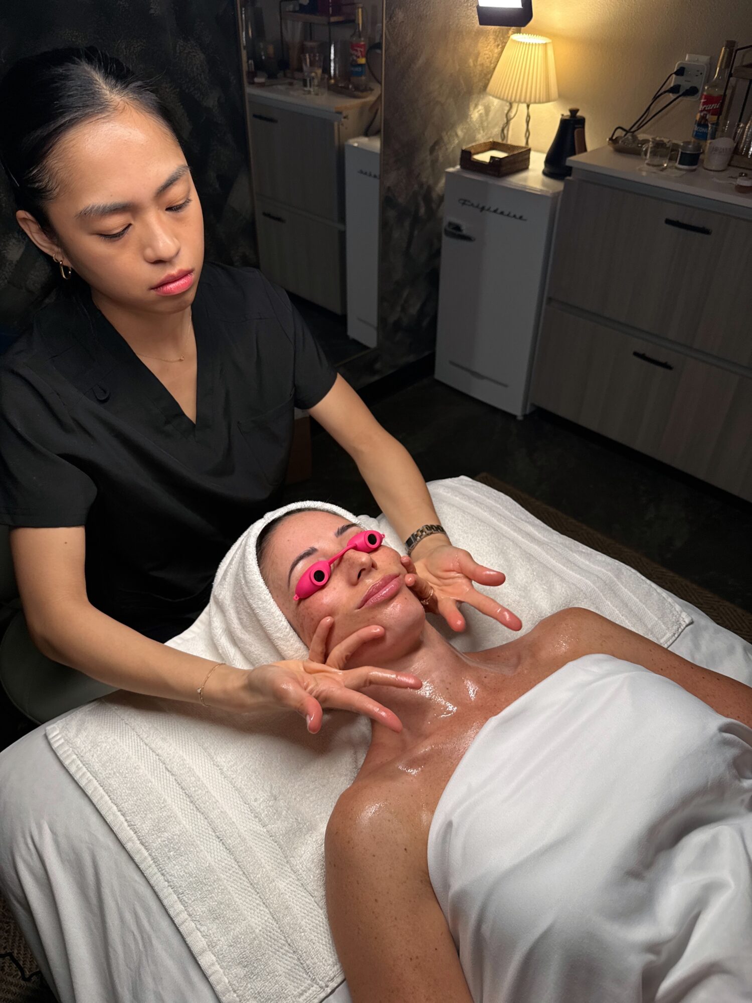 A woman receiving a facial treatment with pink goggles, lying on a bed, with a therapist standing beside her in a spa room.