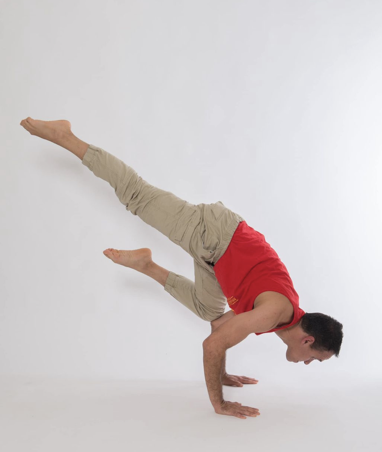 Man balancing on hands with legs extended sideways, wearing a red shirt and beige pants, against a plain white background.