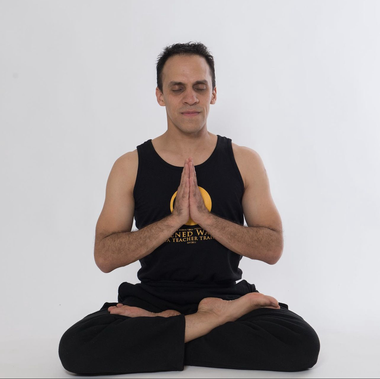Person sitting cross-legged with hands in prayer position, eyes closed, against a plain white background.