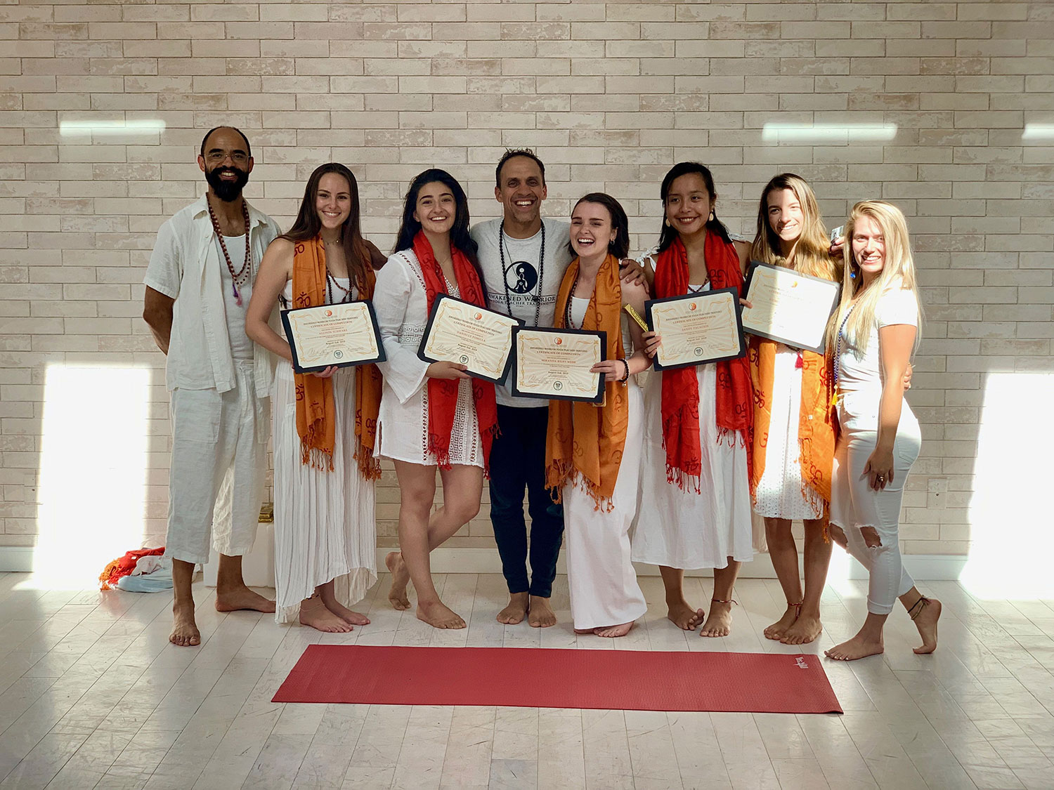 Group of eight people standing on a red carpet holding certificates, smiling, in front of a brick wall.