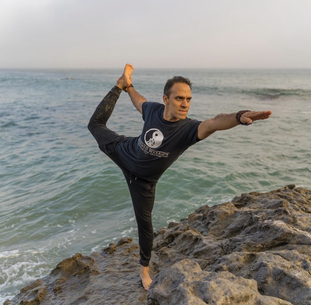 Person practicing yoga on rocks by the sea, balancing on one leg with arms extended, ocean in background.